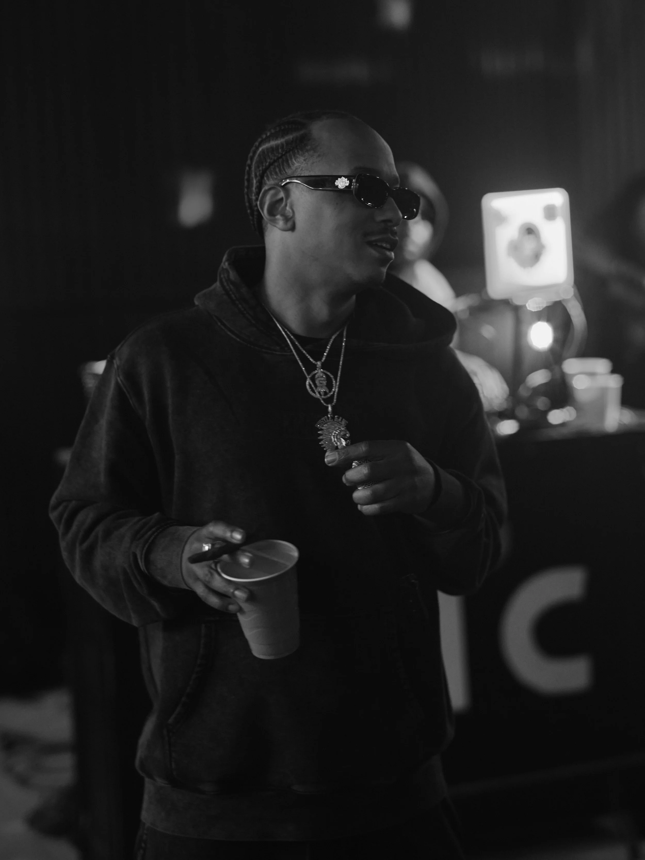 A person wearing dark sunglasses and jewelry, holding a paper cup and a spoon, in a dimly lit environment.