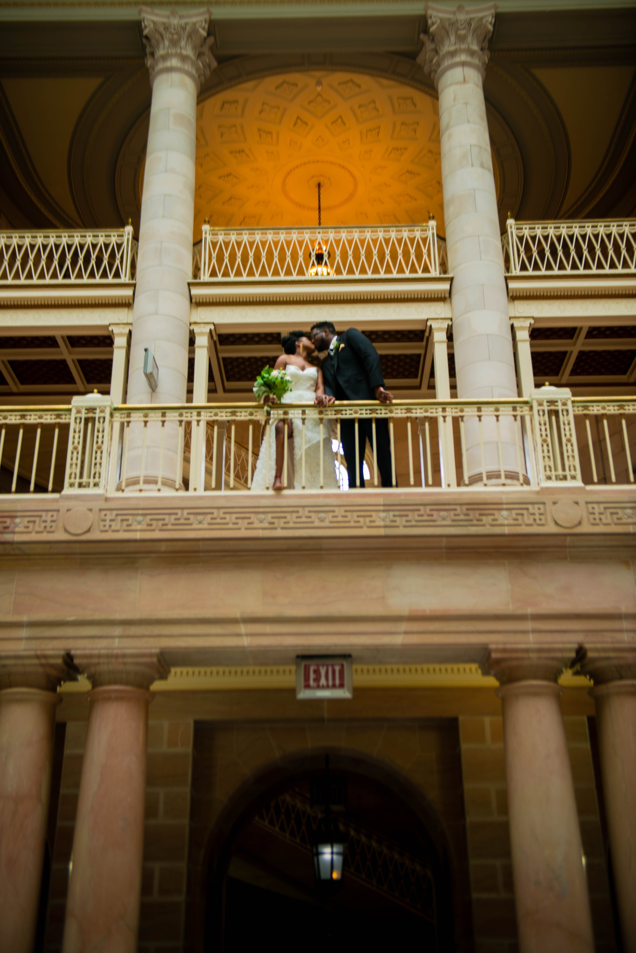 A bride and groom sharing a romantic moment on a balcony of a grand building with tall pillars and ornate architecture.