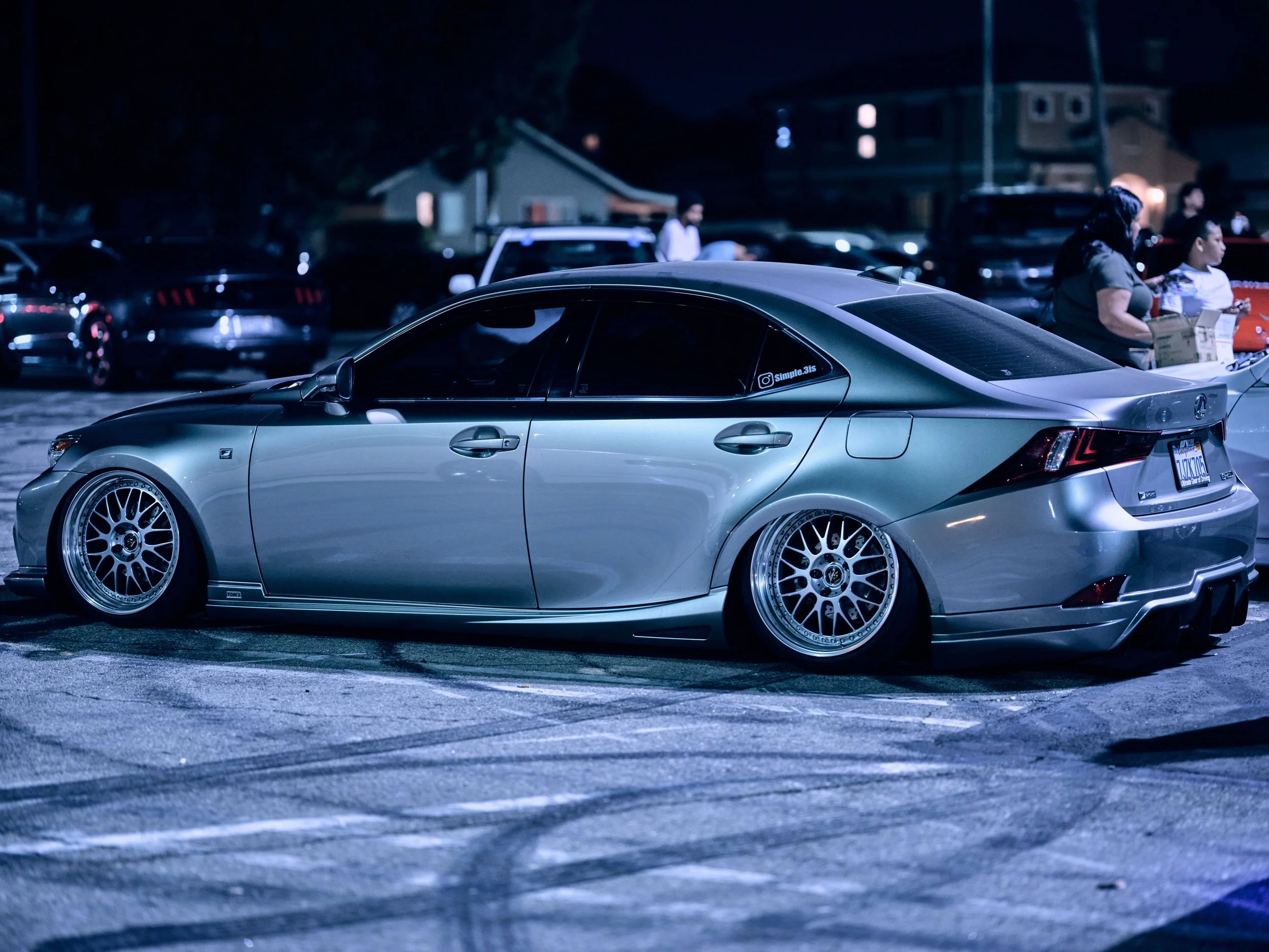 A silver modified Lexus IS car parked in a parking lot at night, with custom wheels, lowered suspension, and a rear diffuser.