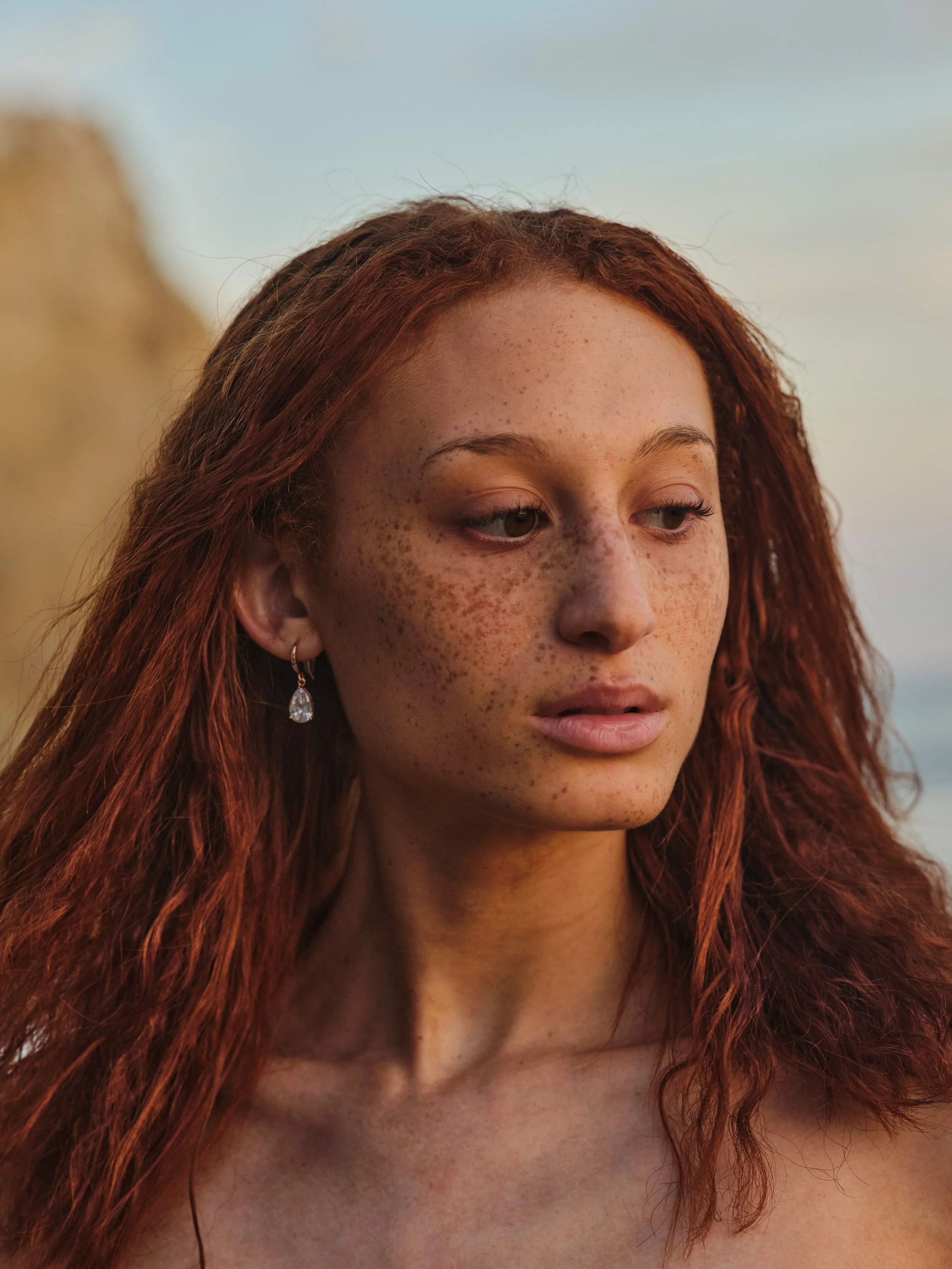 Close-up of a young woman with red hair, freckles, and light makeup, looking contemplatively to the side against a blurred outdoor background.