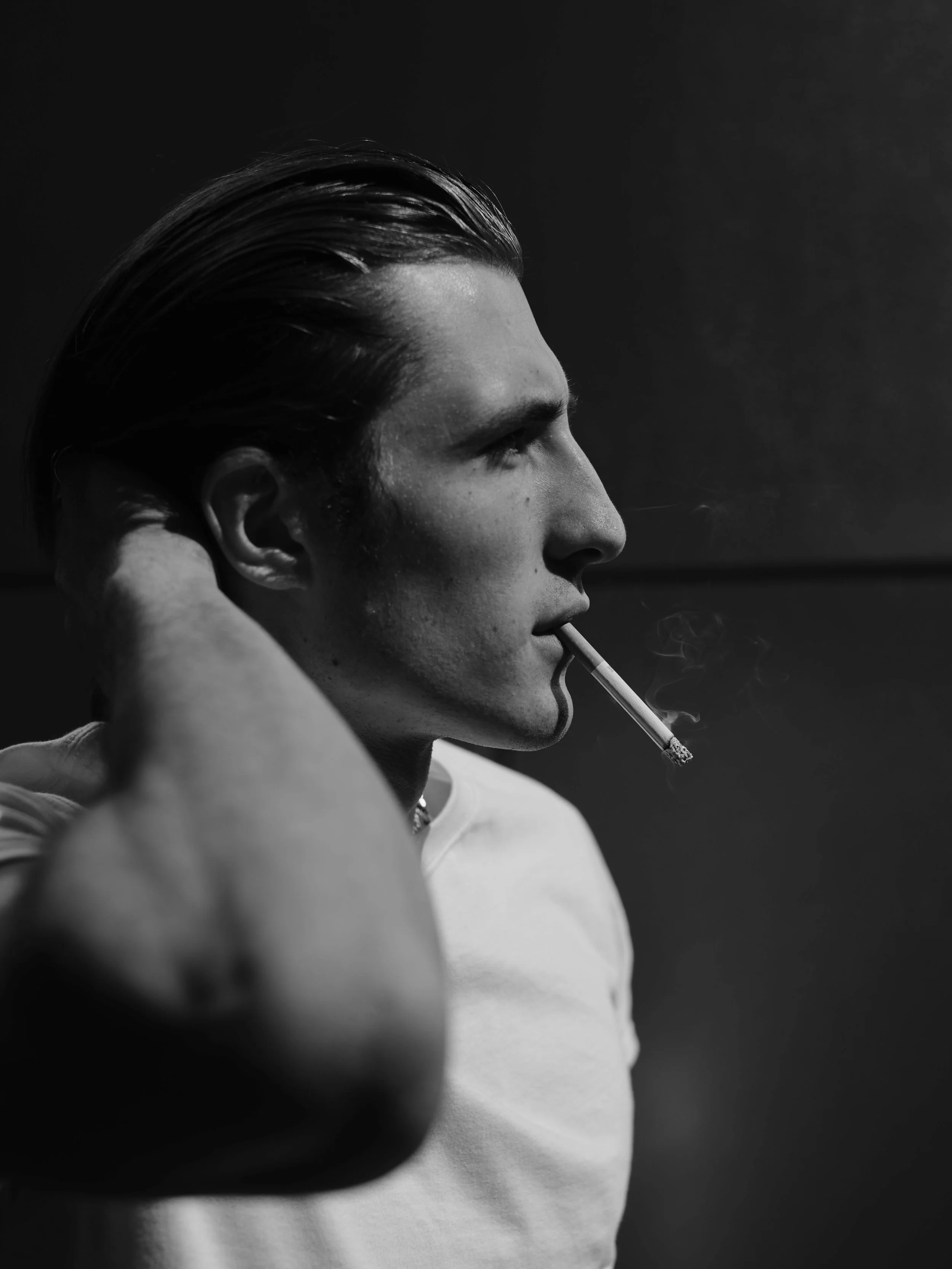 Black and white photo of a man with slicked-back hair smoking a cigarette, with one hand behind his head, wearing a light-colored shirt