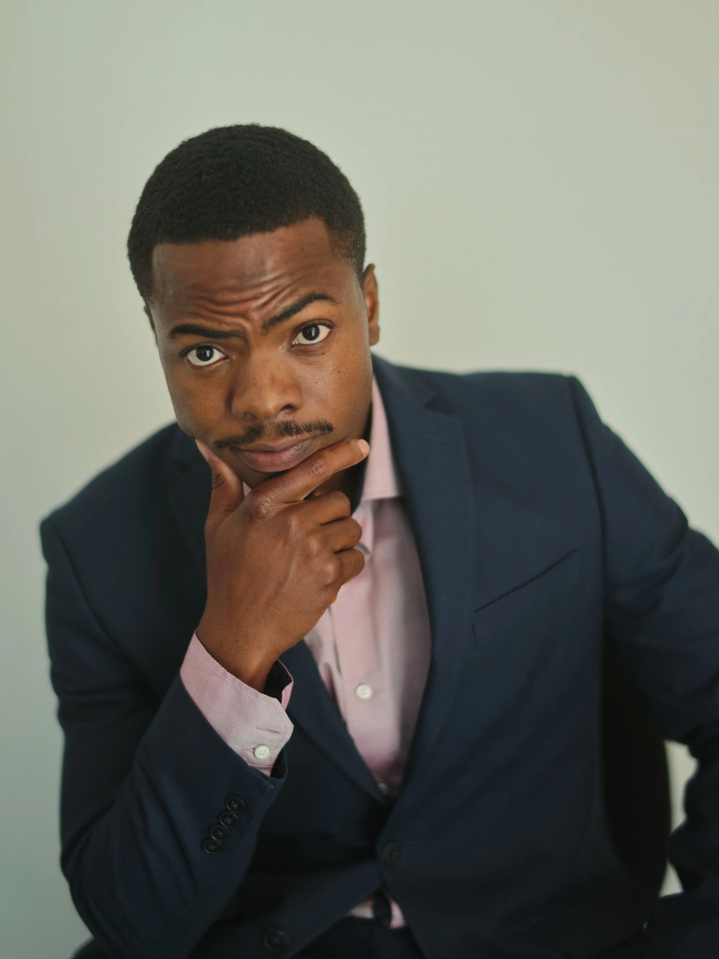 A young man wearing a dark blue suit and pink shirt, sitting with his hand on his chin, looking at the camera with a thoughtful expression.