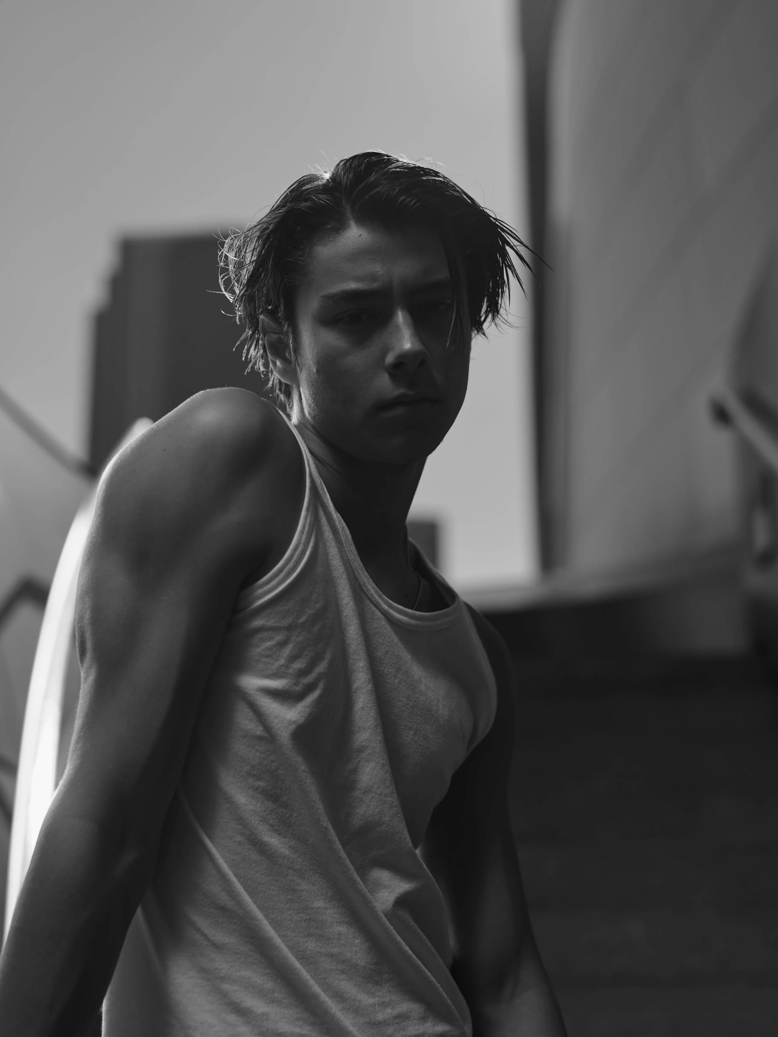 Black and white photo of young person with short, messy hair wearing a tank top, looking intensely at the camera, with urban background.