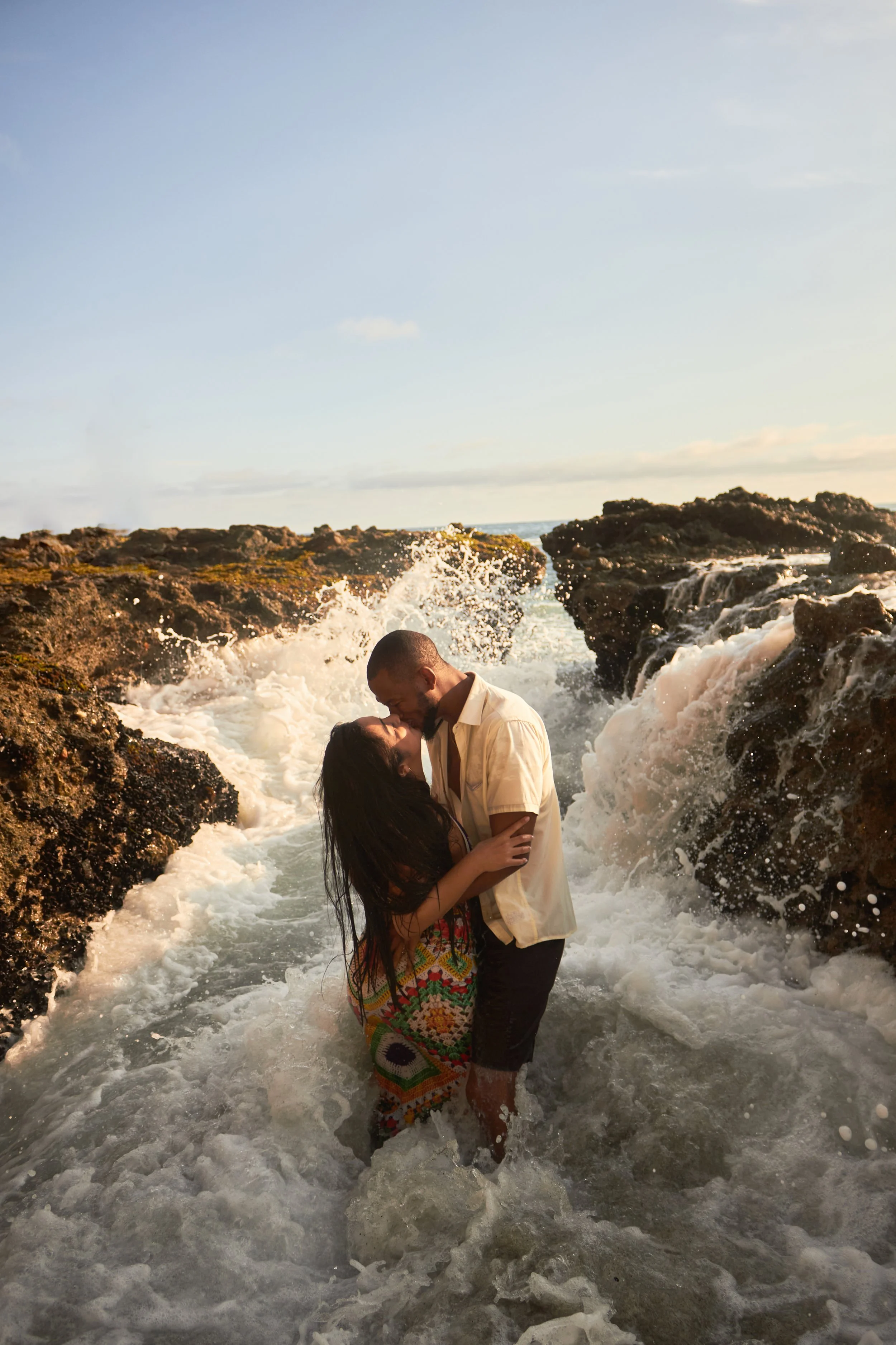 A couple is kissing in the ocean waters near rocky shoreline, with waves splashing around them during sunset.