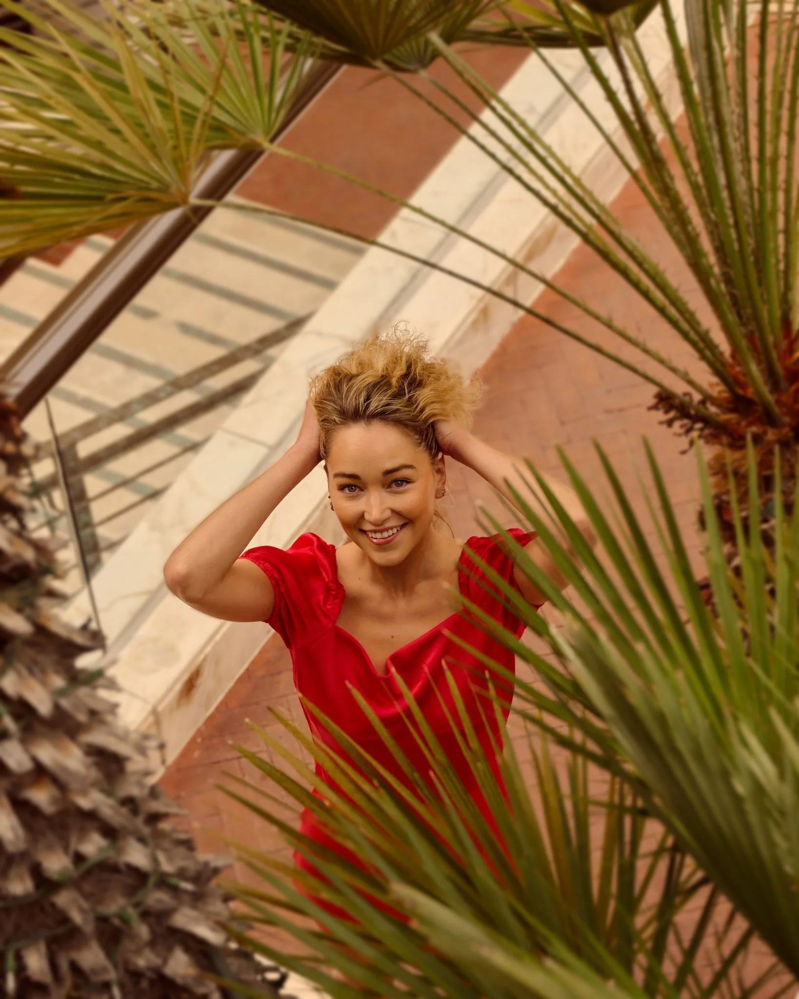 A smiling woman wearing a red dress, looking up at the camera, surrounded by green palm leaves, on a balcony with a metal railing.