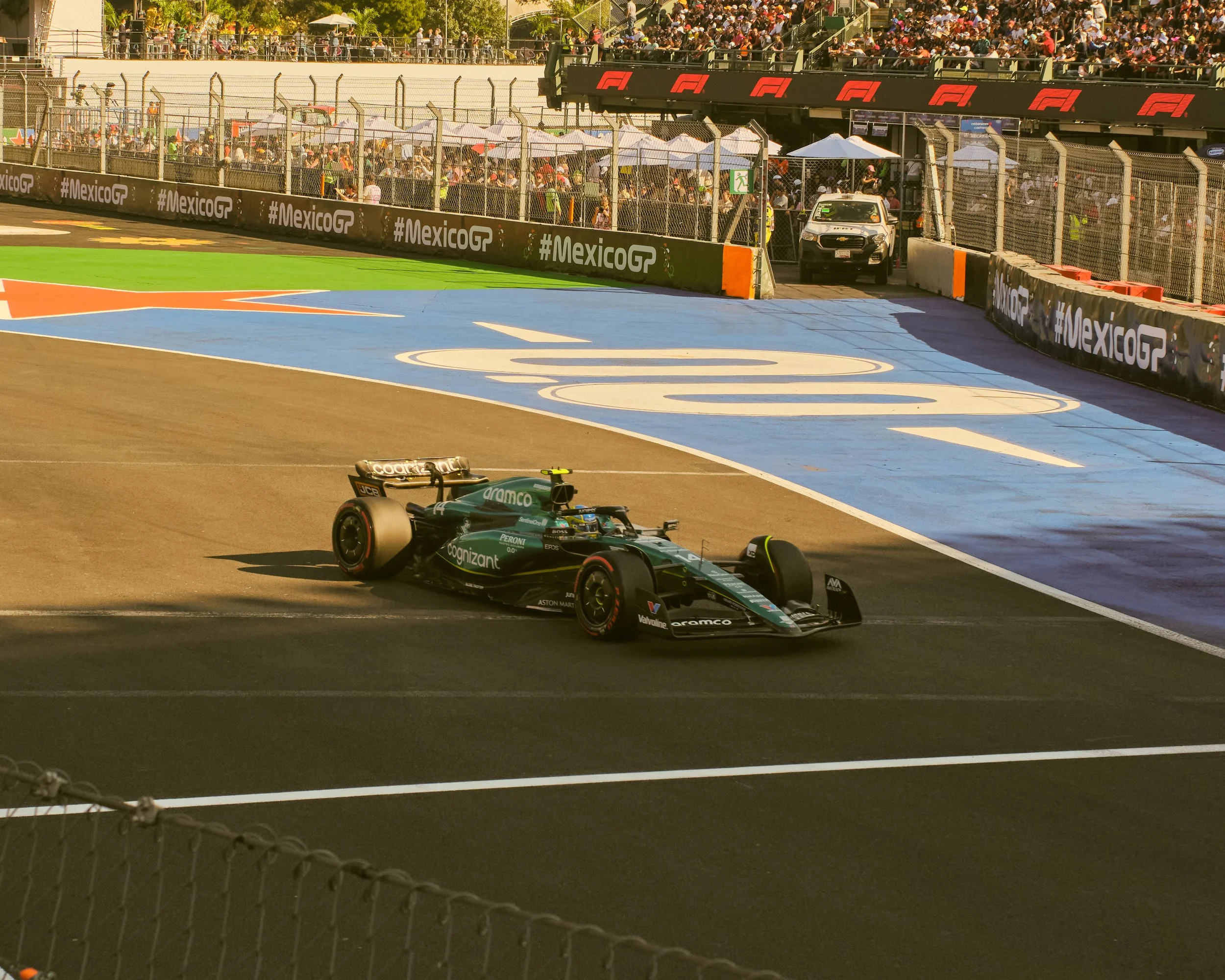 A race car on a racing track during a Formula 1 race at the Mexico City Grand Prix with spectators in the stands and a car parked near the edge of the track.