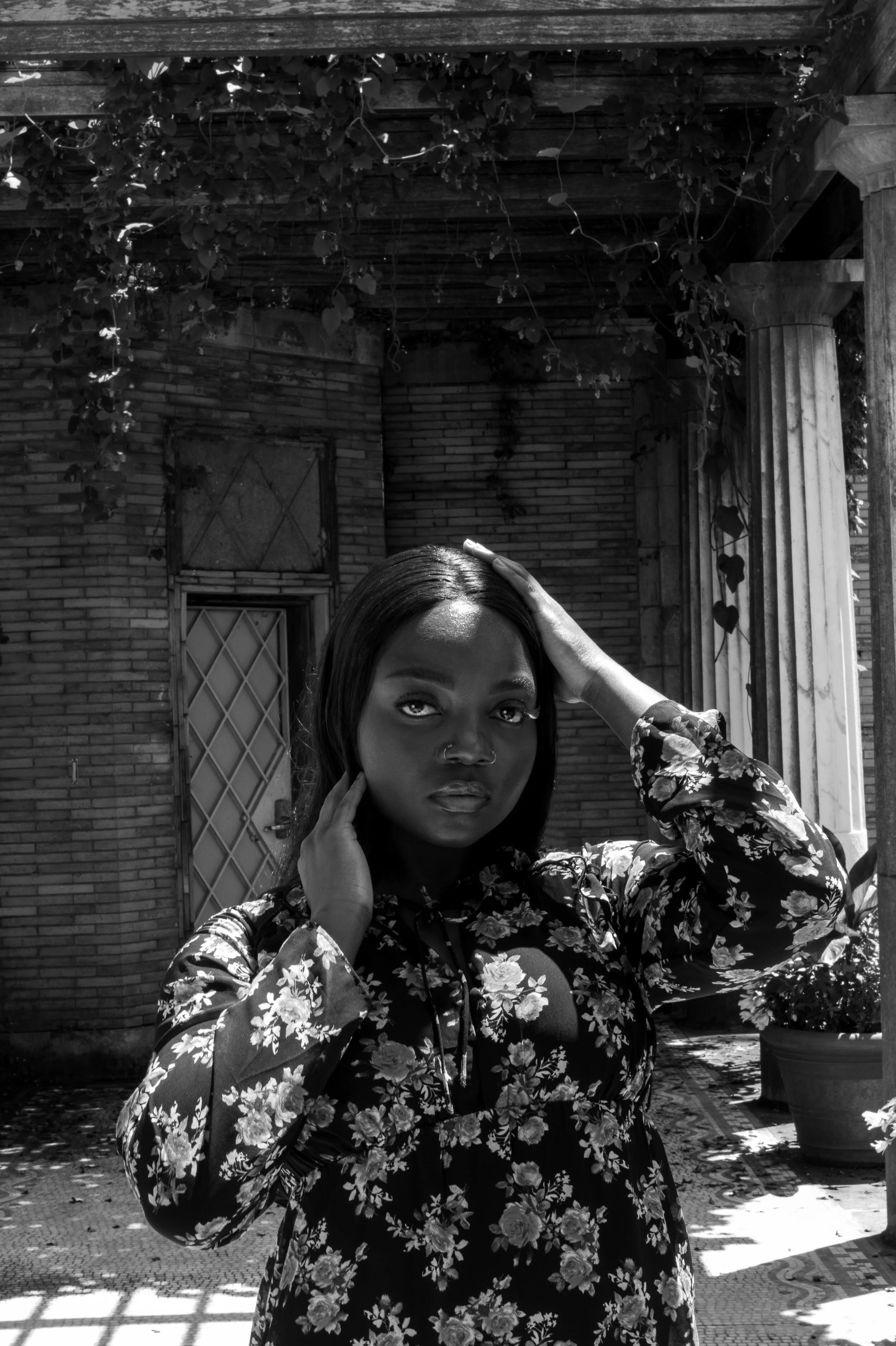 A woman with dark hair, wearing a floral dress, posing outdoors with her hand on her head and a serious expression, near brick walls and greenery.