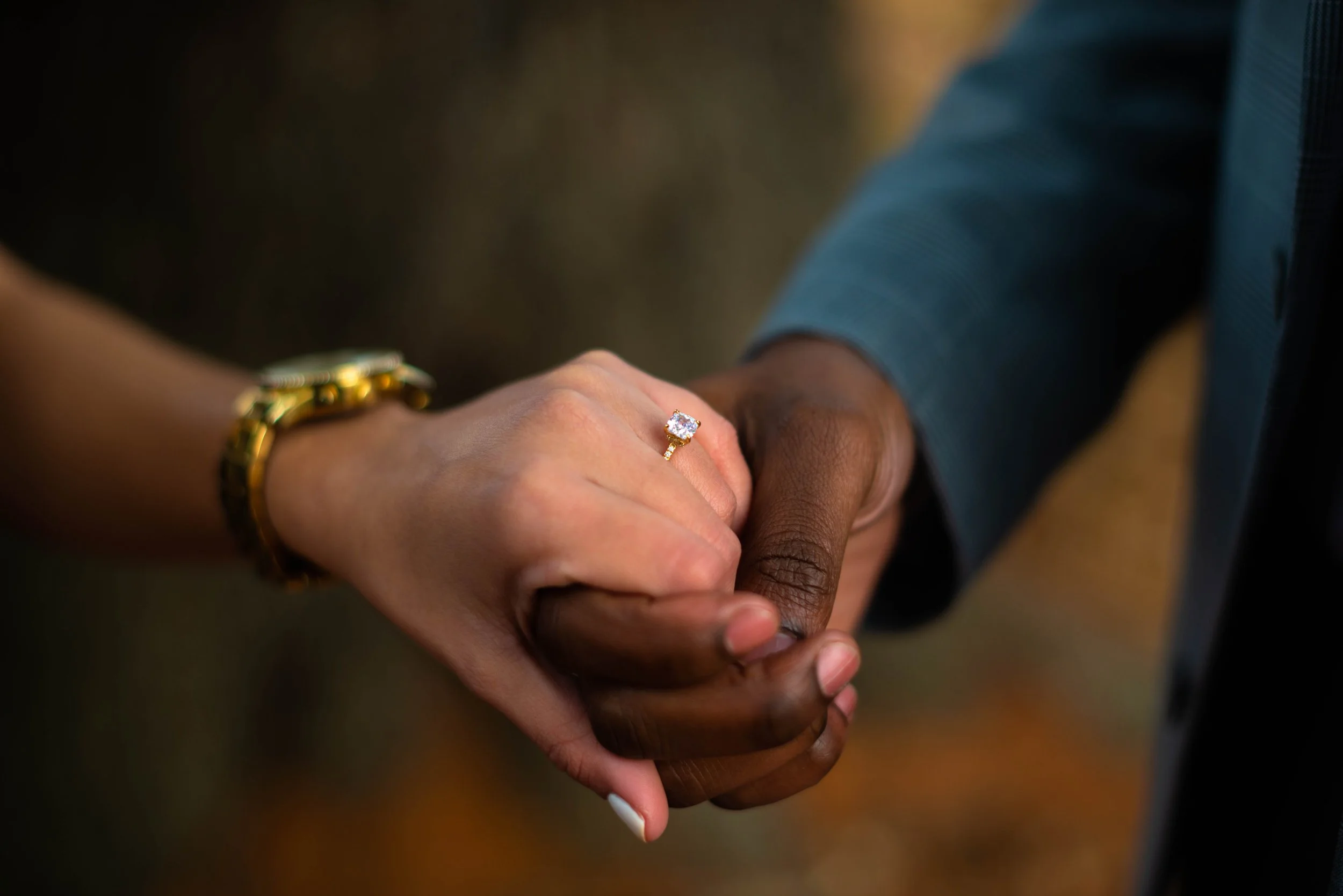 Close-up of a couple holding hands, showcasing an engagement ring with a large diamond on a woman's finger. The woman wears a gold watch and has a manicured nail, while the man is dressed in a formal suit with a blue shirt.