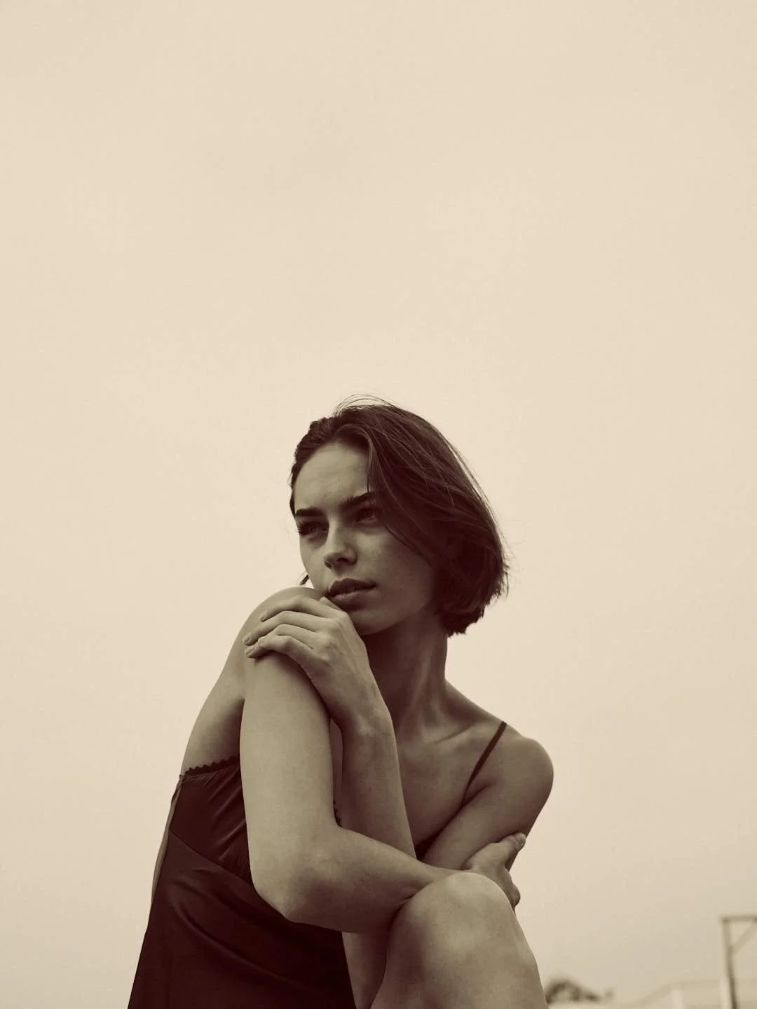 A young woman with short dark hair, wearing a spaghetti strap top, sitting with one knee up, hand resting on her shoulder, looking contemplative against a plain, beige background.