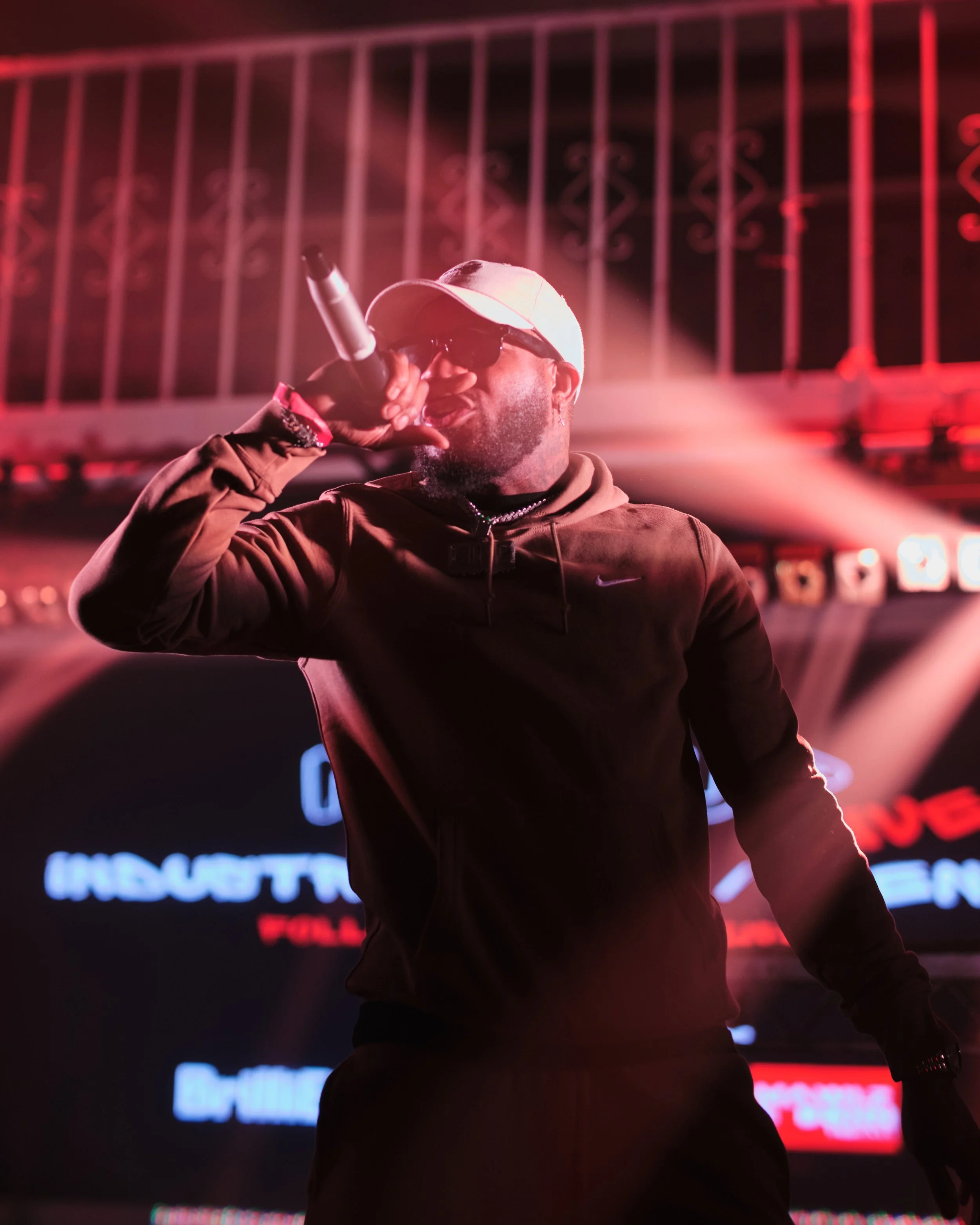 A man wearing sunglasses and a baseball cap holding a microphone and singing on stage with red and white lighting.