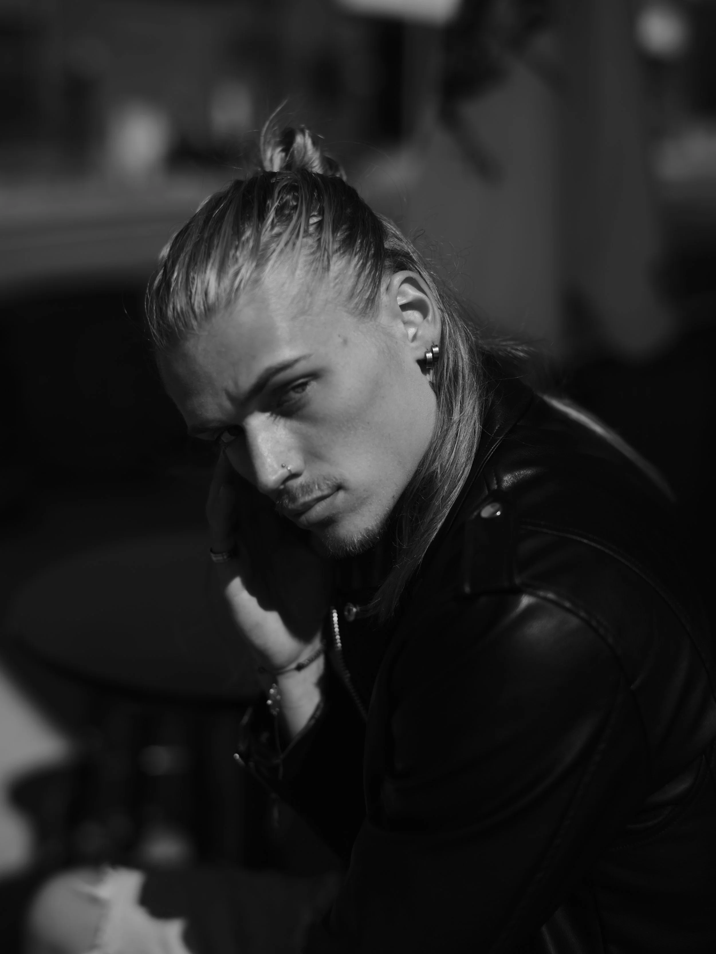 Black and white photo of a young man with long hair, styled in a top knot, wearing earrings and a leather jacket, looking intensely at the camera.