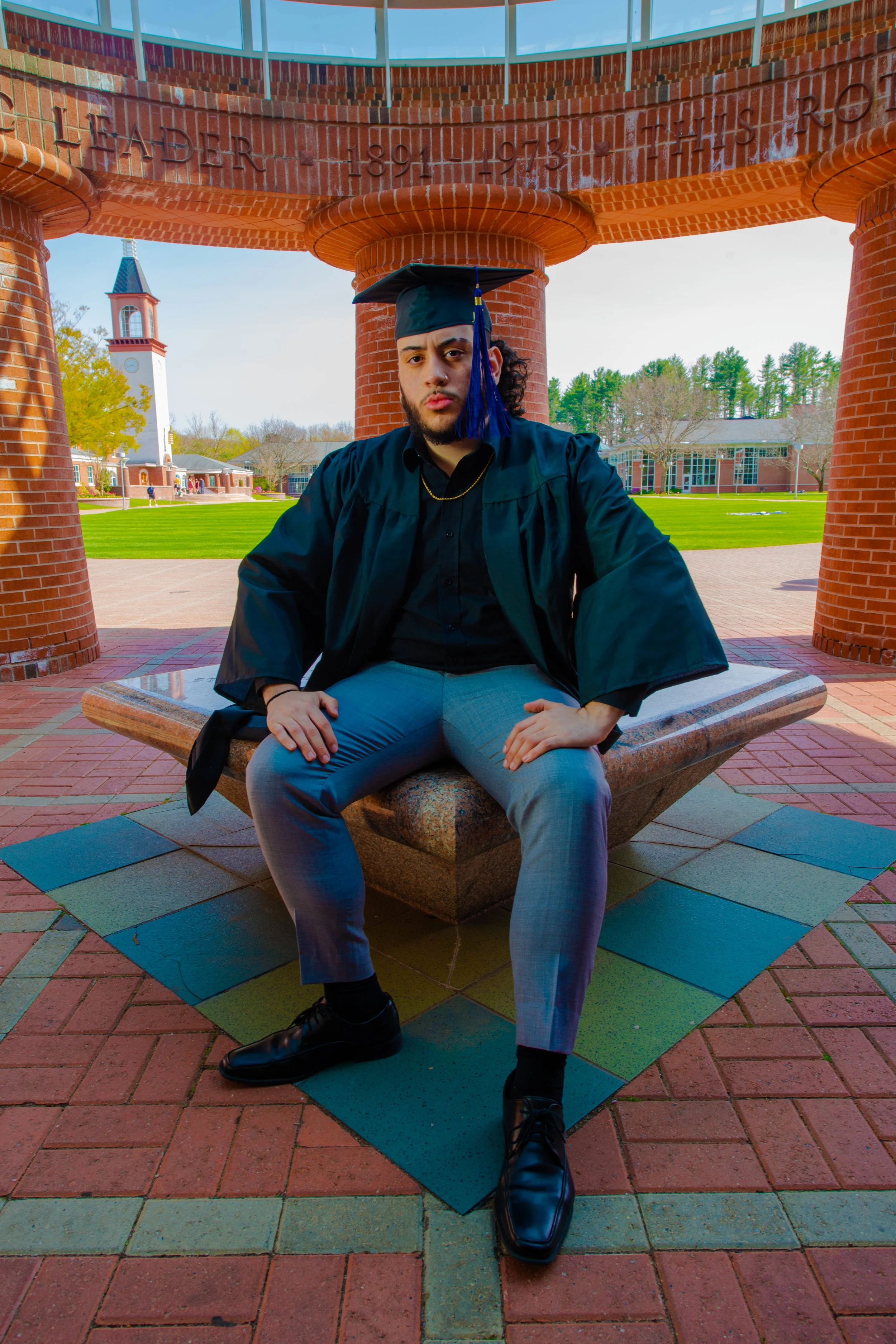 A young man in a graduation cap and gown sitting on a stone bench in an outdoor area with a campus in the background.