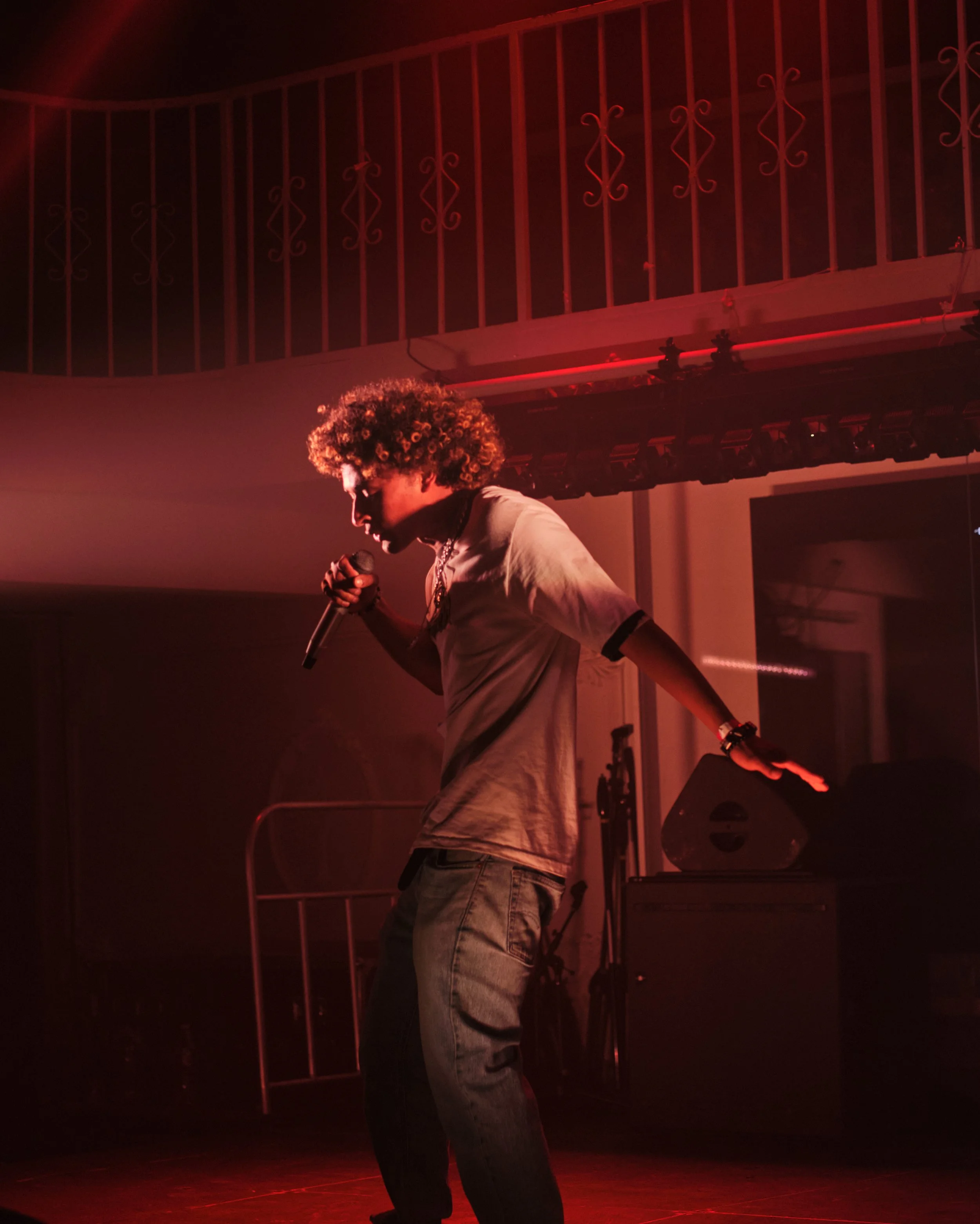 A person with curly hair singing into a microphone on stage, illuminated by red lighting.