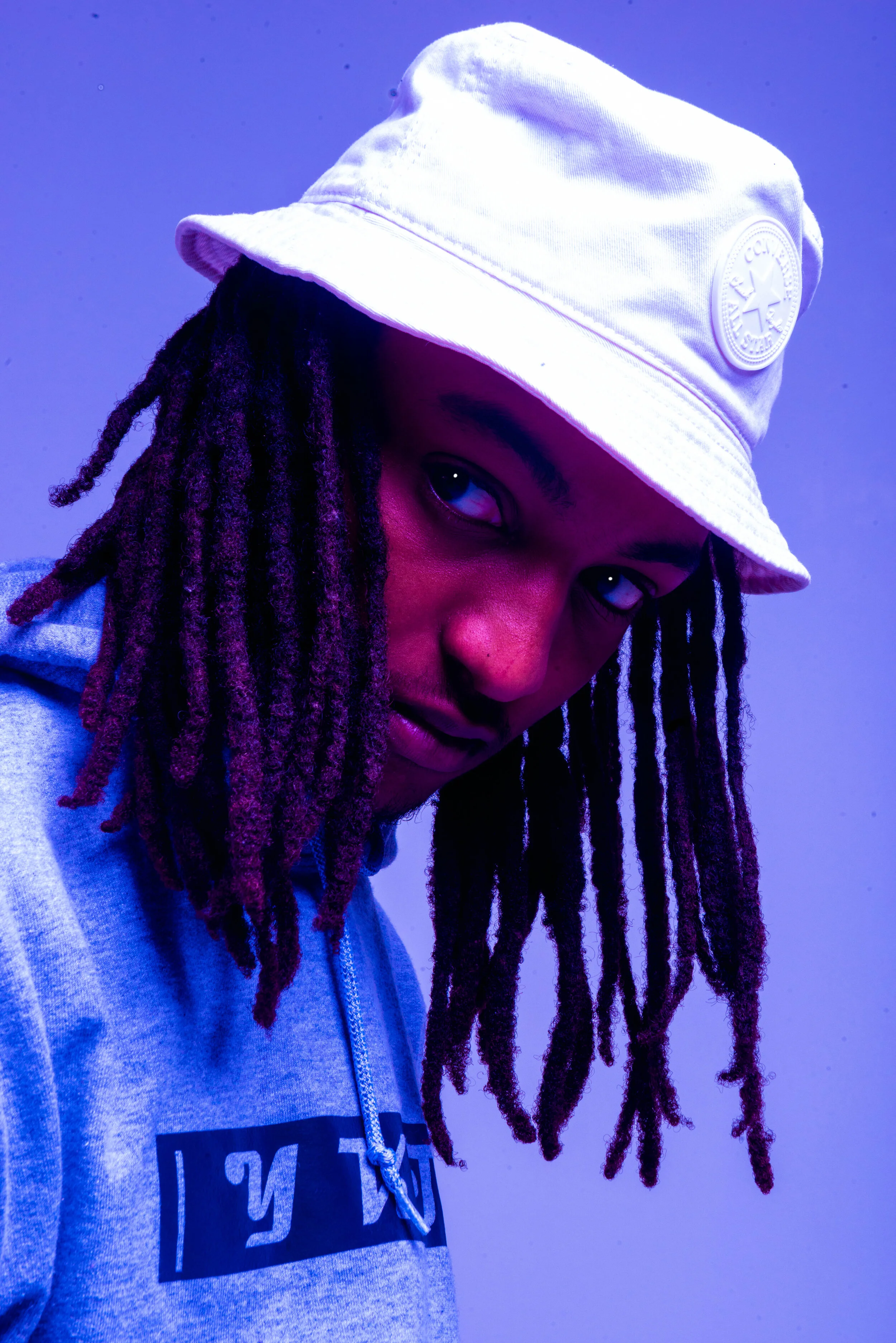 A person with long dreadlocks wearing a white bucket hat and a grey hoodie, looking over their shoulder against a blue background.