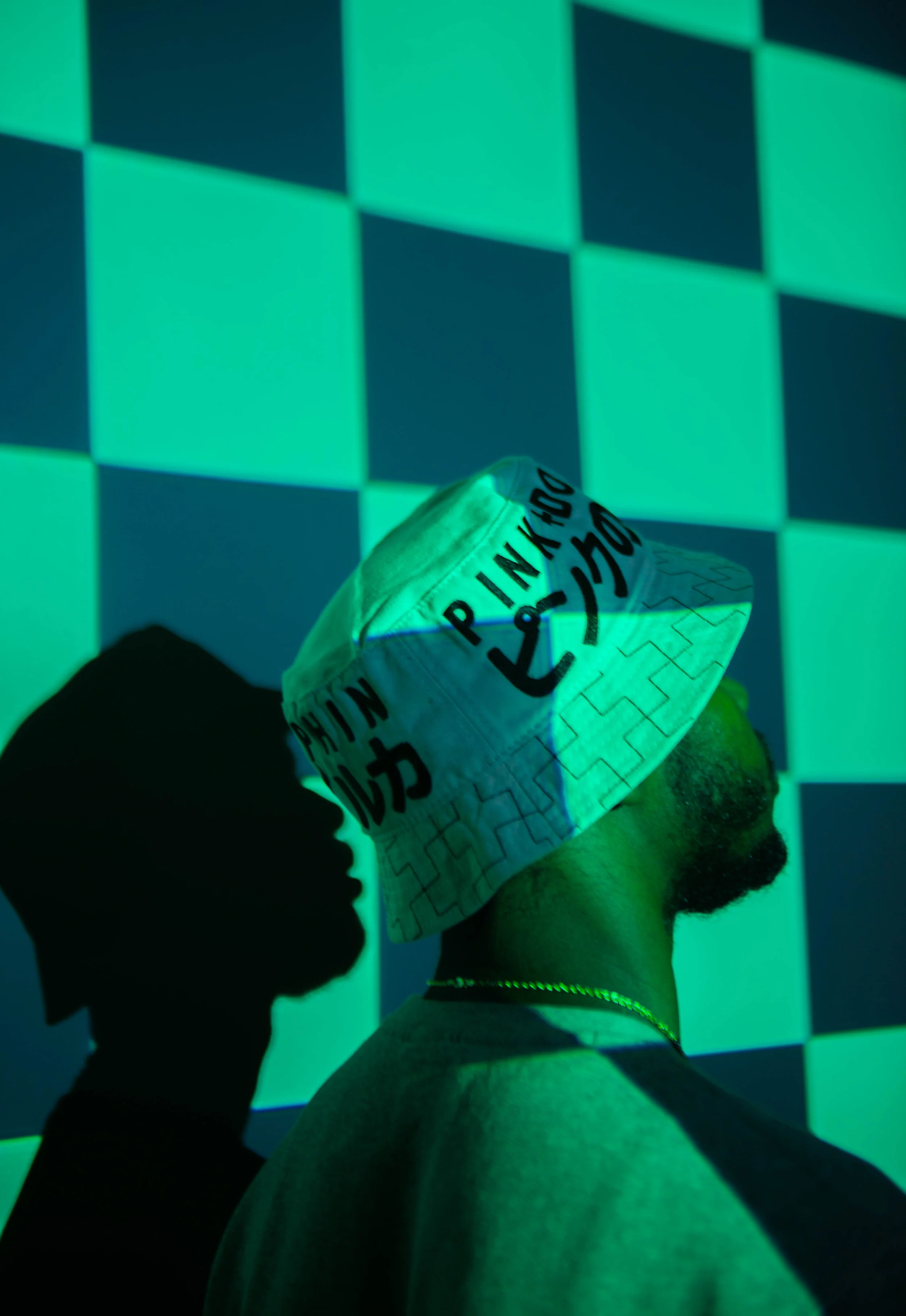 Person wearing a white bucket hat with black text, shadow of a person's face on the wall, background of glowing green and blue checkered pattern.