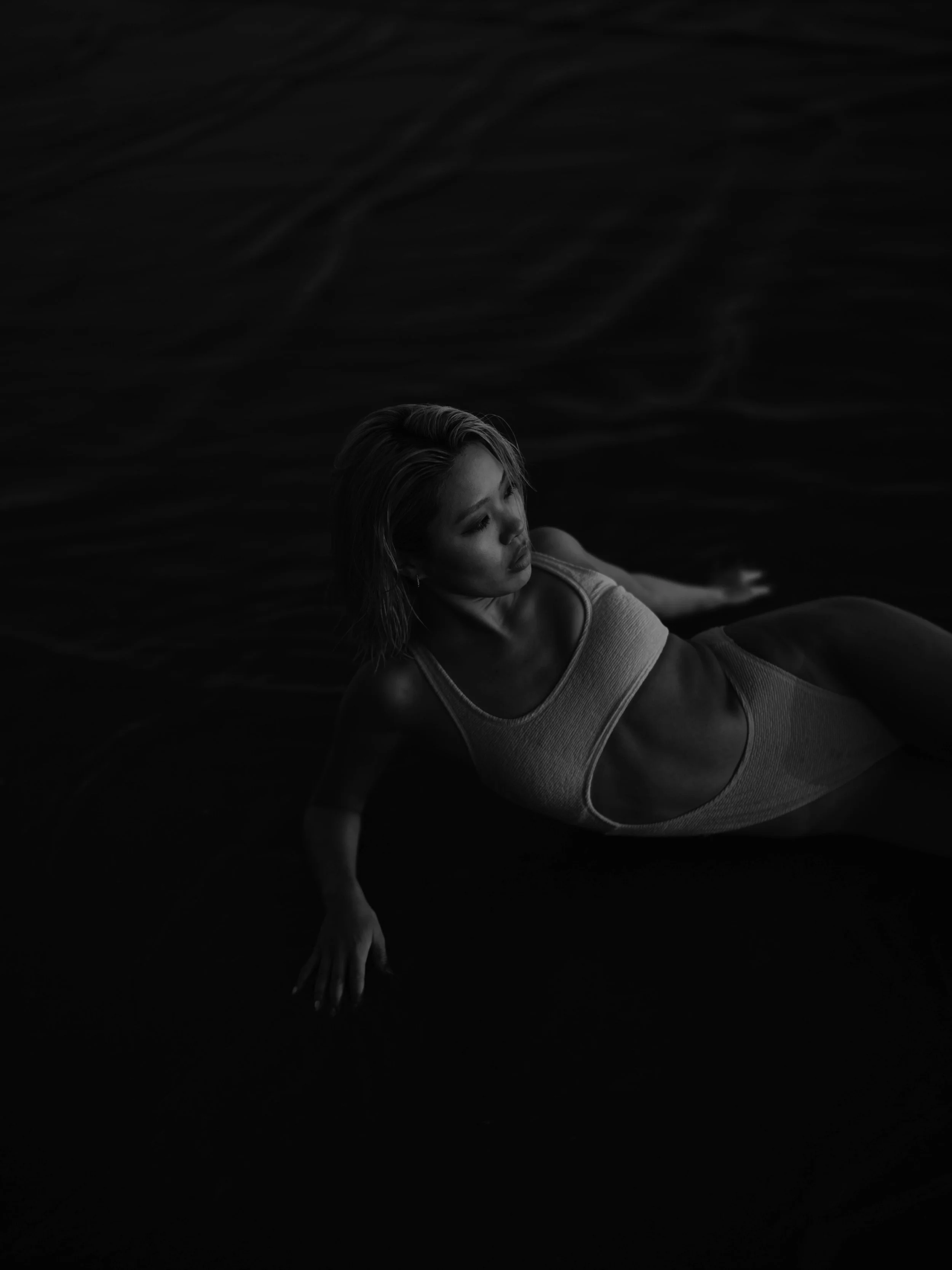 A woman in a one-piece swimsuit lying on her side in dark water, looking contemplative.