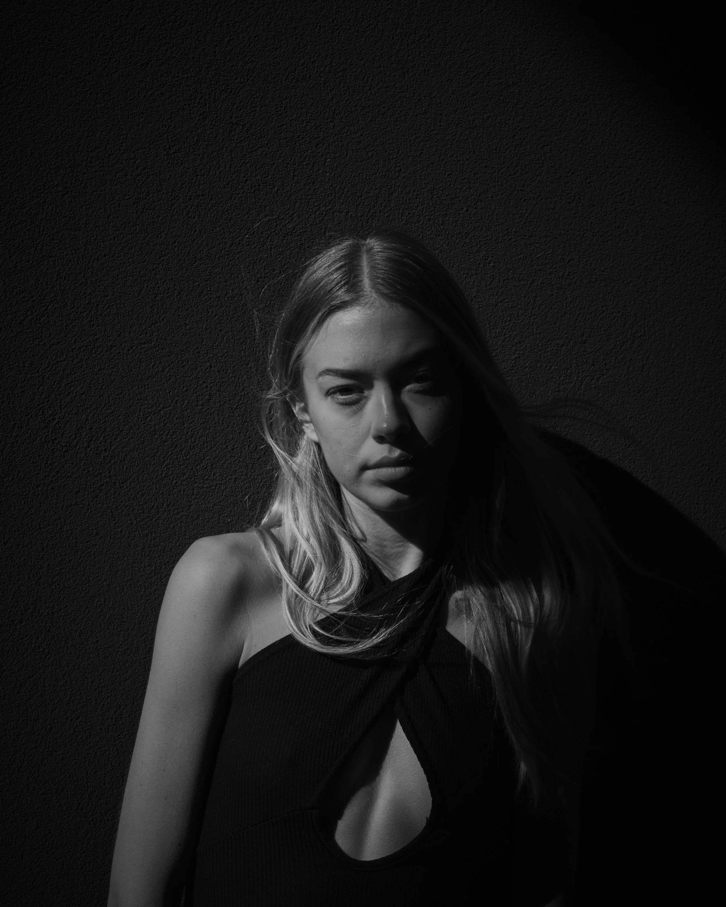A black and white portrait of a young woman with long hair, standing against a dark textured wall, with shadows casting across her face and body.