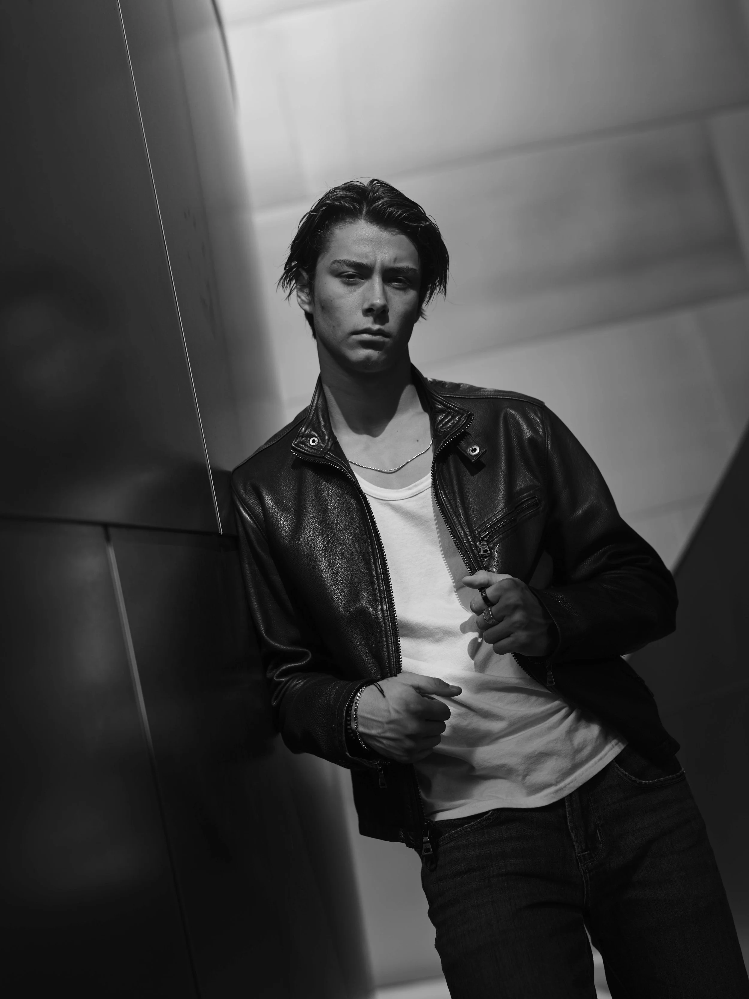 A young man with short hair, wearing a leather jacket, white T-shirt, and jeans, leaning against a metallic wall in a modern indoor setting, looking serious.
