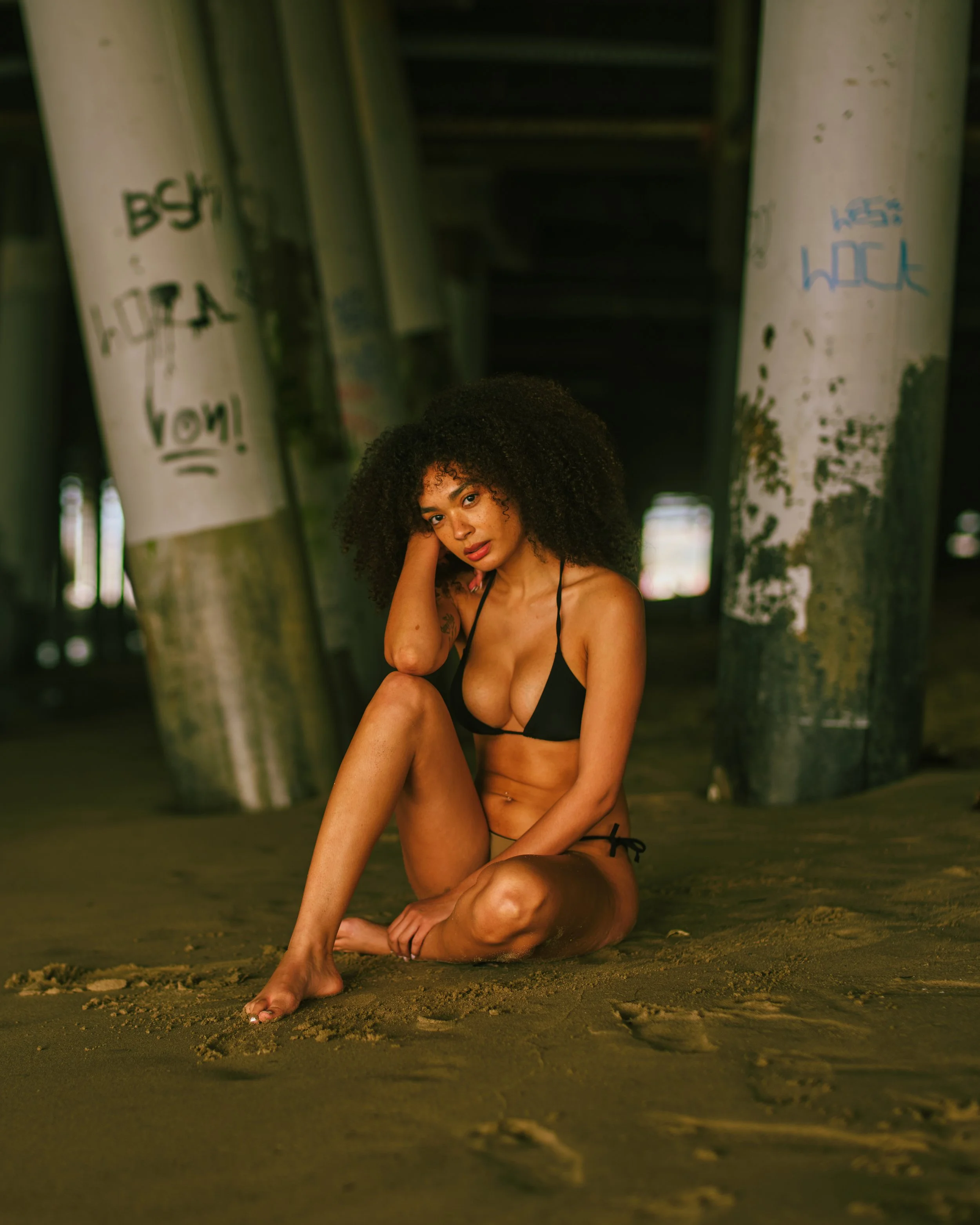 A woman with curly hair wearing a black bikini sitting on sandy ground under a bridge with graffiti-covered pillars.