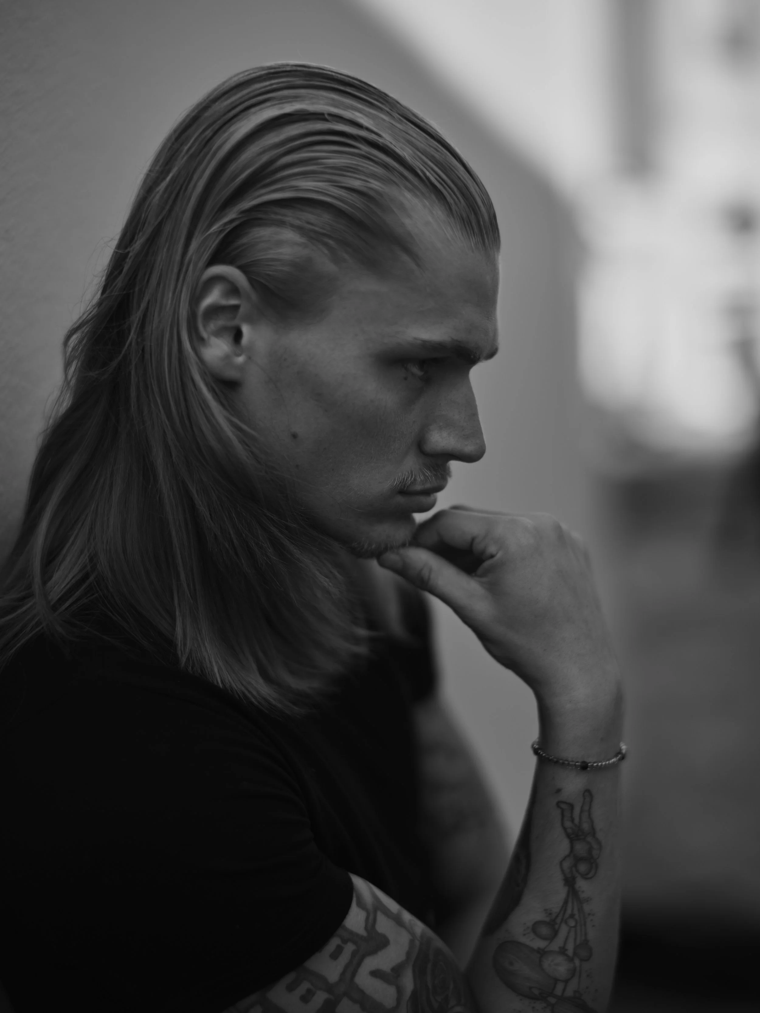 Black and white portrait of a young man with long, slicked-back hair and tattoos on his arm, with his hand resting on his chin and looking down thoughtfully.