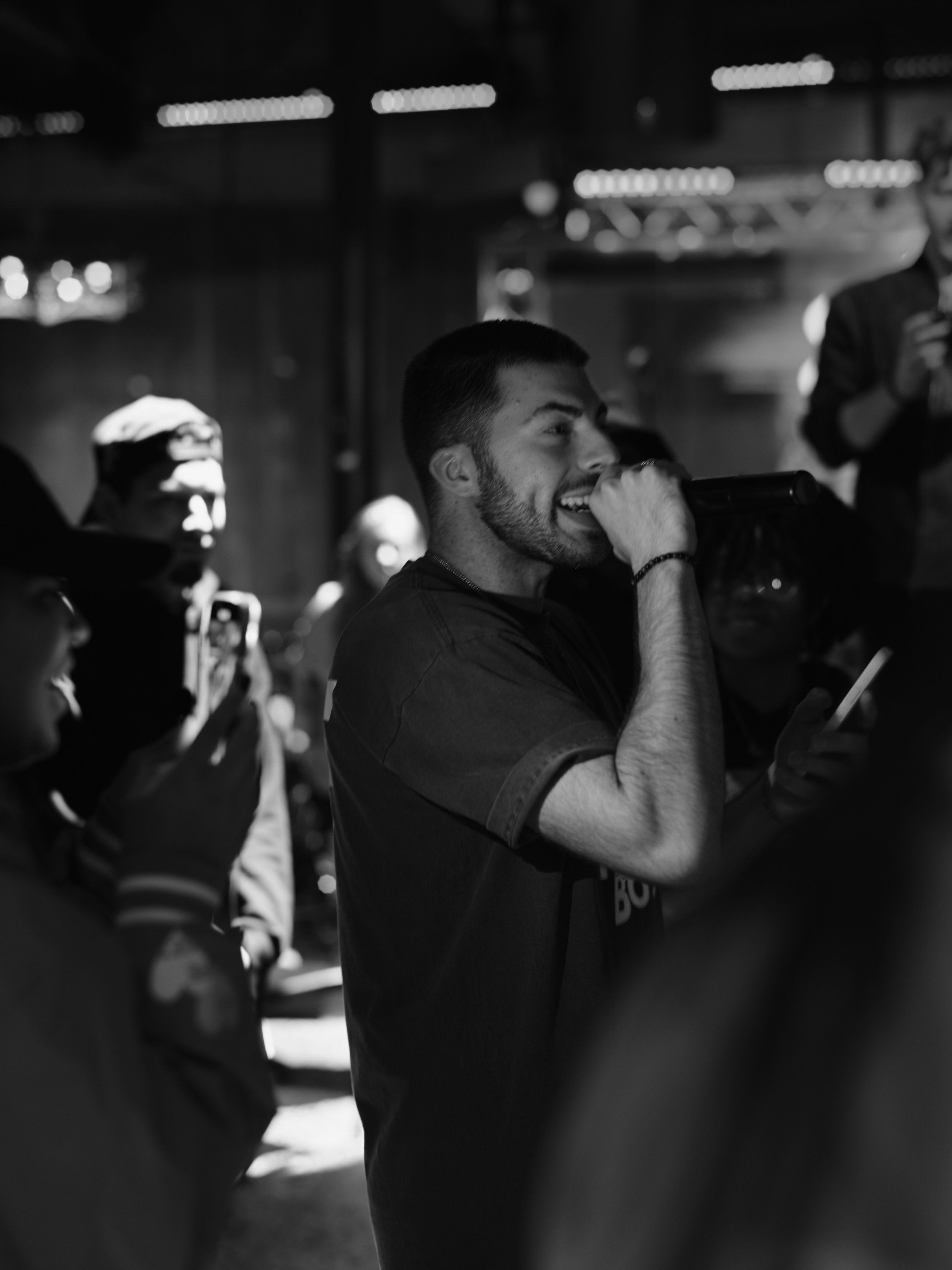 Young man singing into a microphone at an indoor event or concert, surrounded by other people including a woman taking a photo, in black and white.