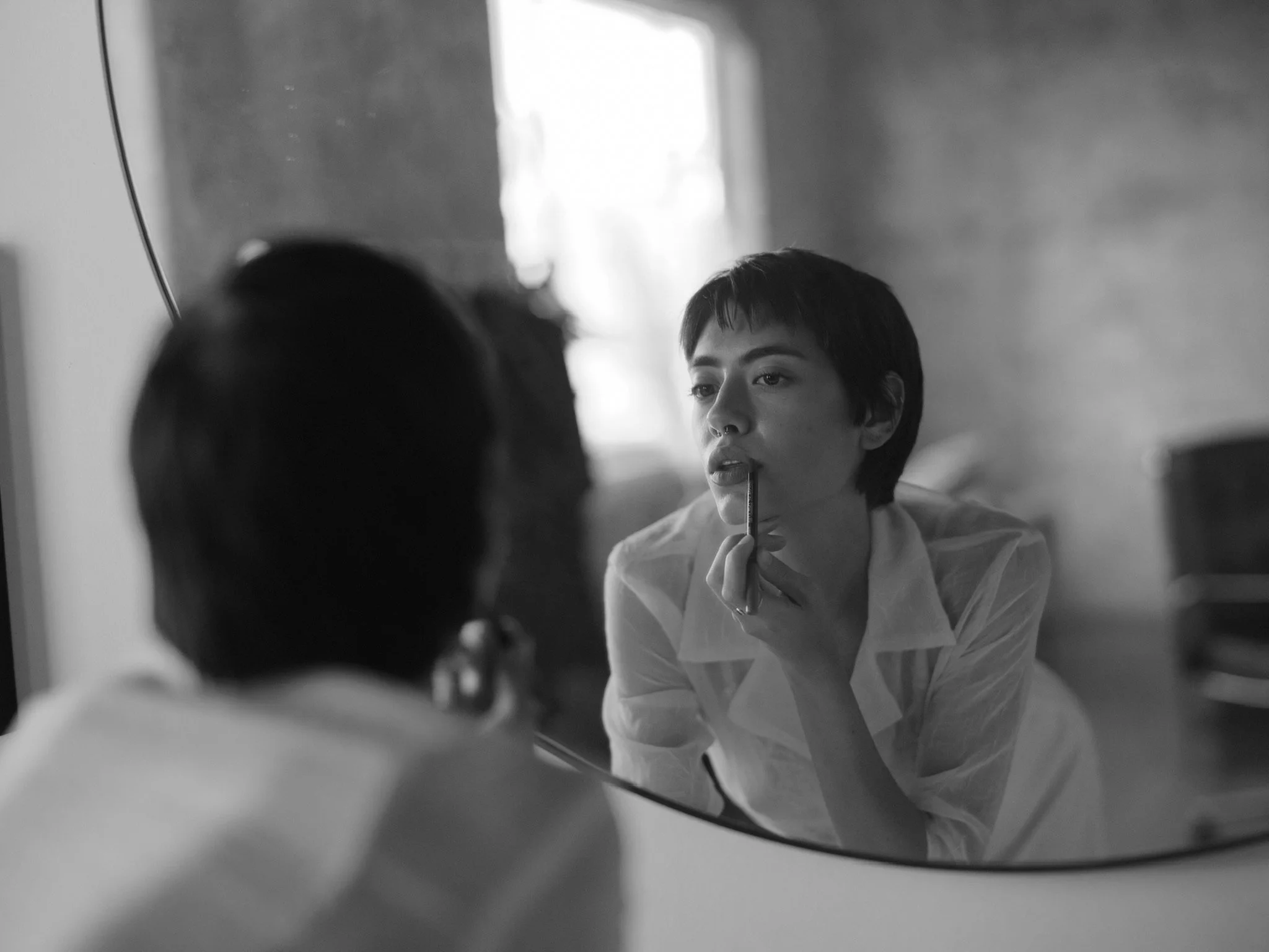 A woman with short hair putting on makeup in front of a mirror, seen from behind.