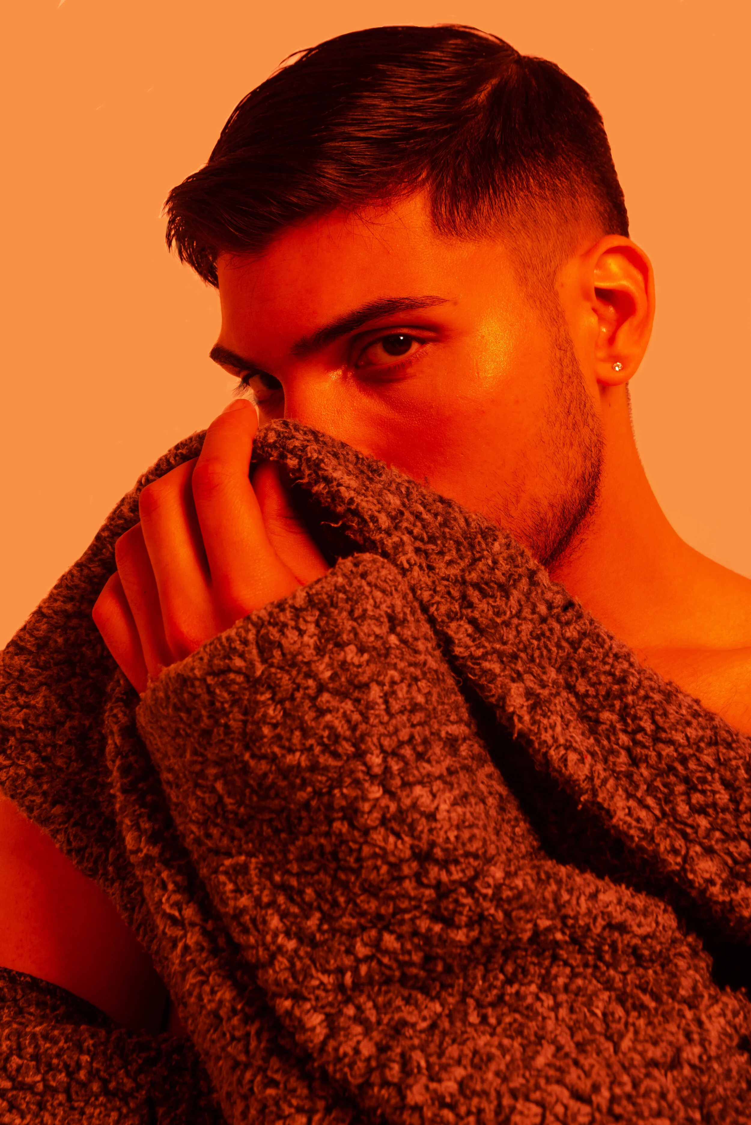 A young man with short dark hair, wearing earrings, partially covering his face with a brown fuzzy coat, looking directly at the camera against a warm orange background.