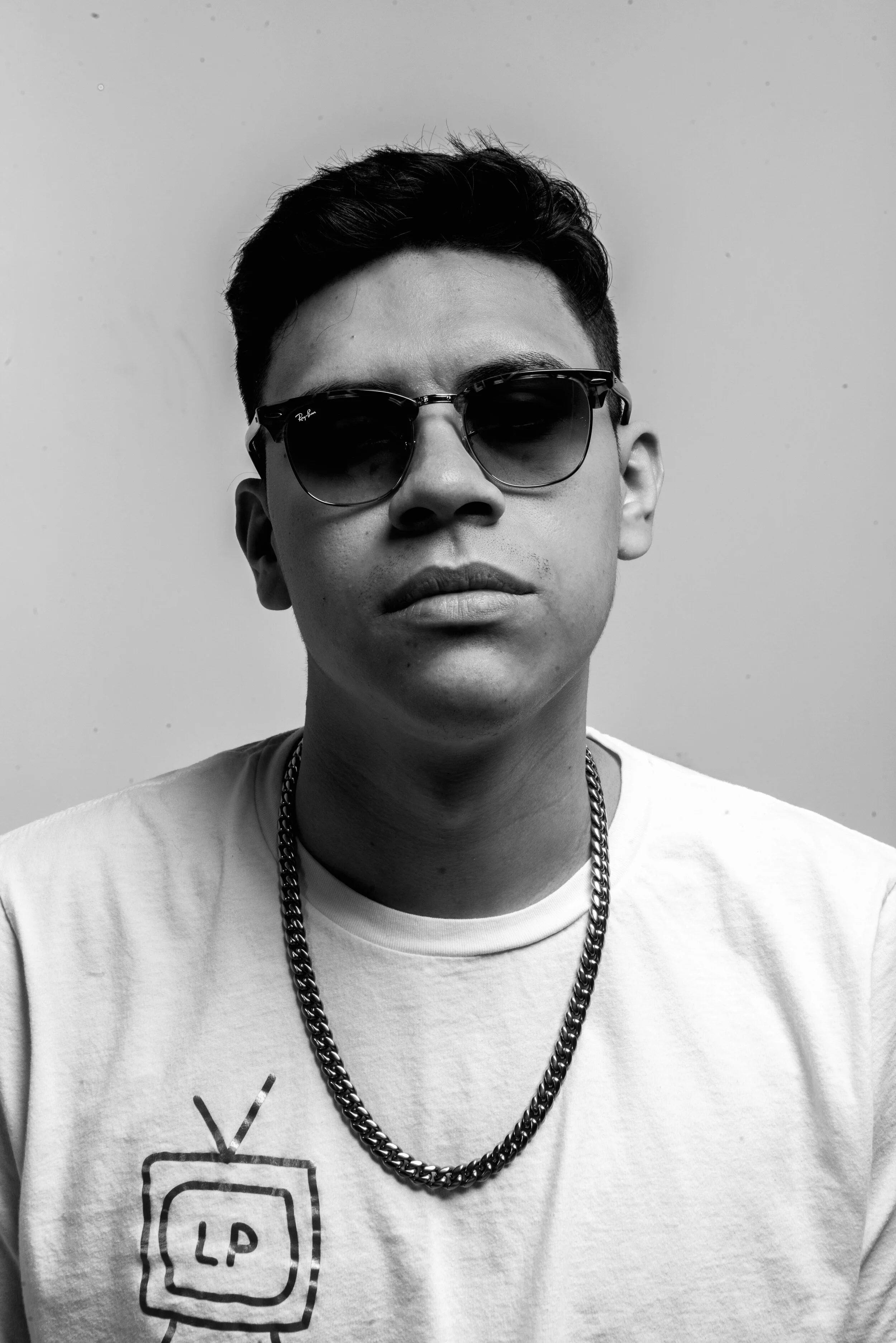 A young man wearing sunglasses and a white t-shirt with a small graphic related to television. He has short, dark hair and is wearing a chain necklace. The background is plain and light-colored.