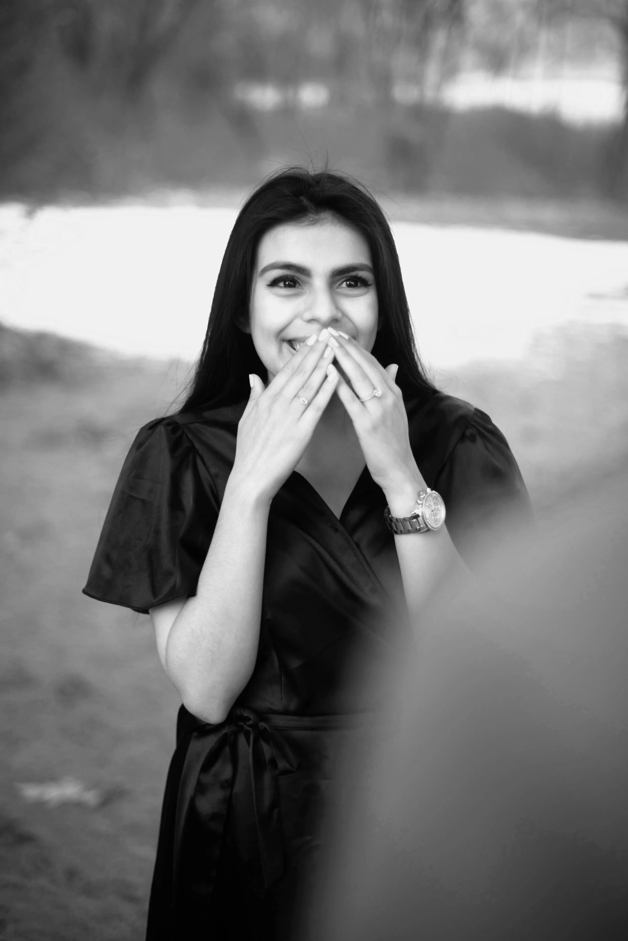 A young woman with long dark hair, smiling, covering her mouth with her hands, wearing a watch and rings, standing outdoors by a lake or pond, in a black-and-white photo.