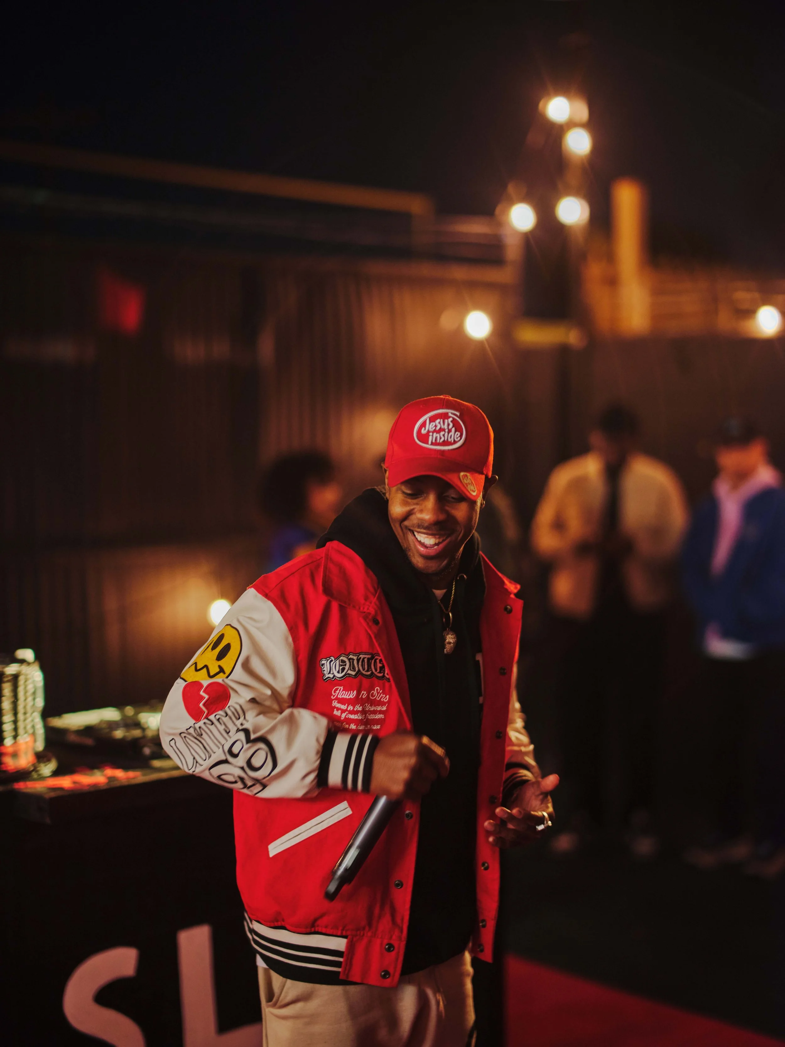 A smiling man wearing a red jacket and red cap with 'Jesus Inside' written on it, holding a microphone, at a party or event with people in the background.