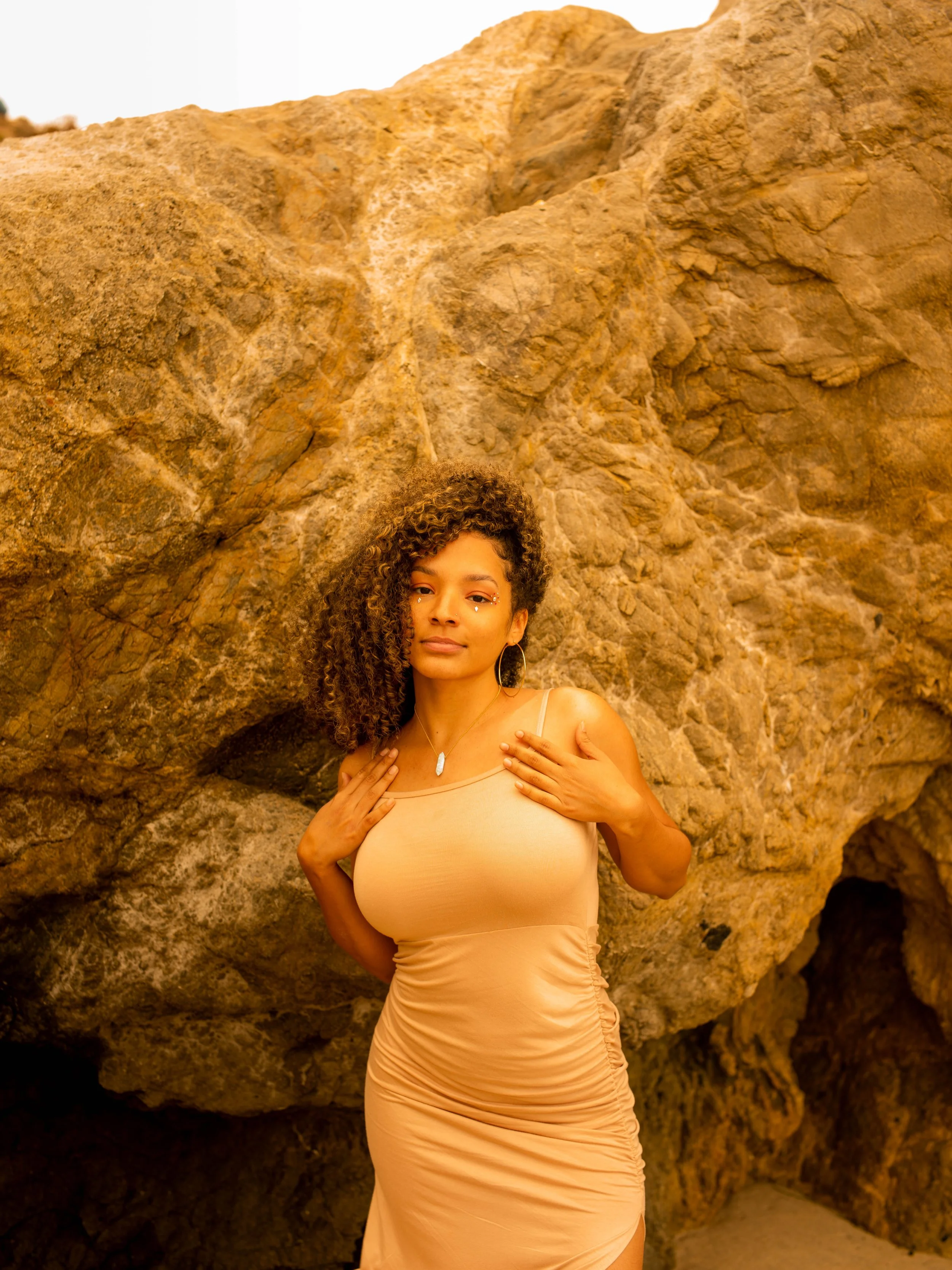 A woman with curly hair standing in front of large rock formations on a desert landscape. She is wearing a beige, form-fitting dress and jewelry, looking at the camera with her hands touching her chest.