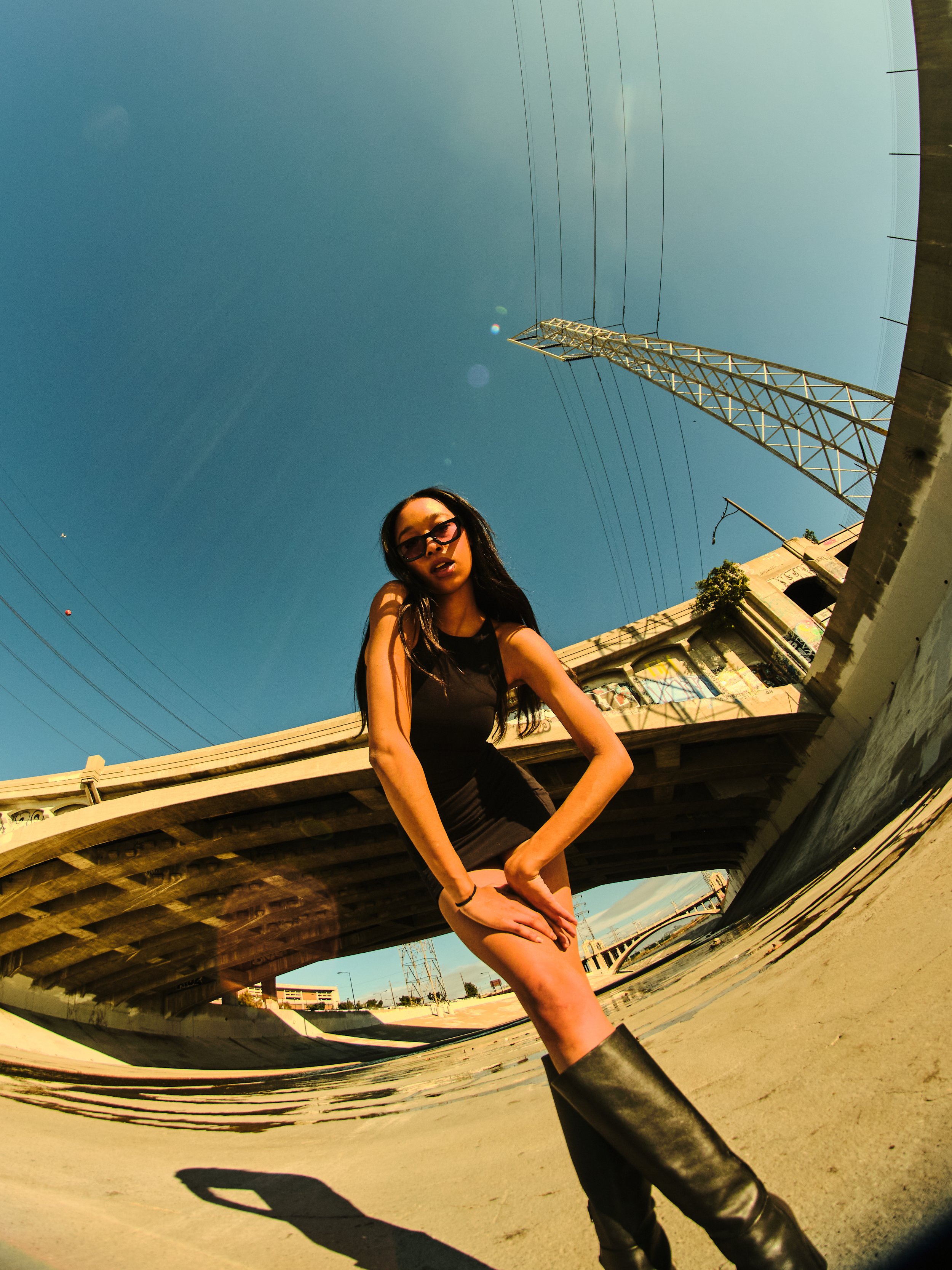 A young woman with long dark hair and glasses, wearing a black sleeveless top, standing outdoors under a clear blue sky near a bridge and electrical tower.