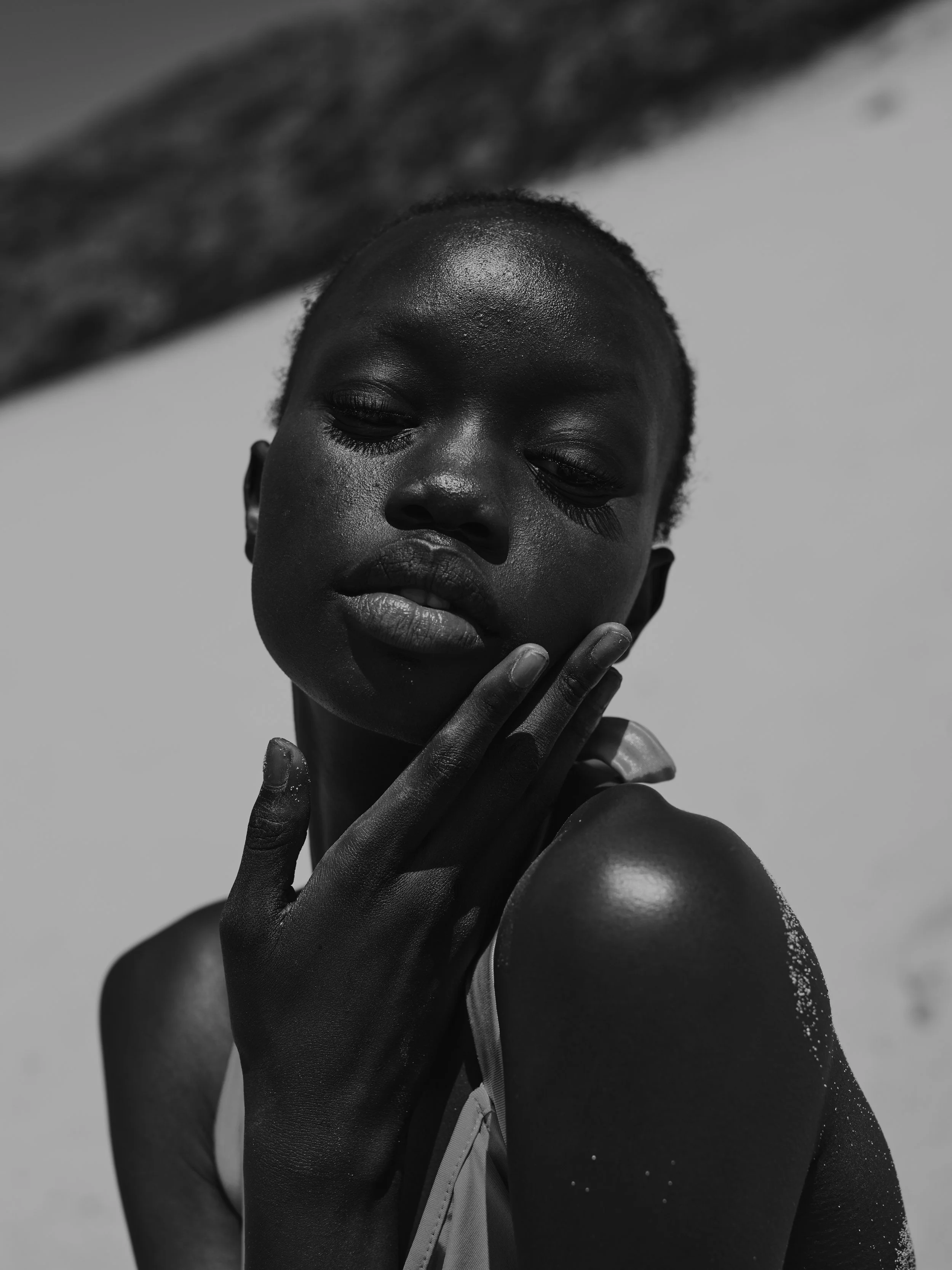 Close-up black and white portrait of a young woman with short hair, looking downward with her hand gently touching her face and shoulder, against a bright sky background.