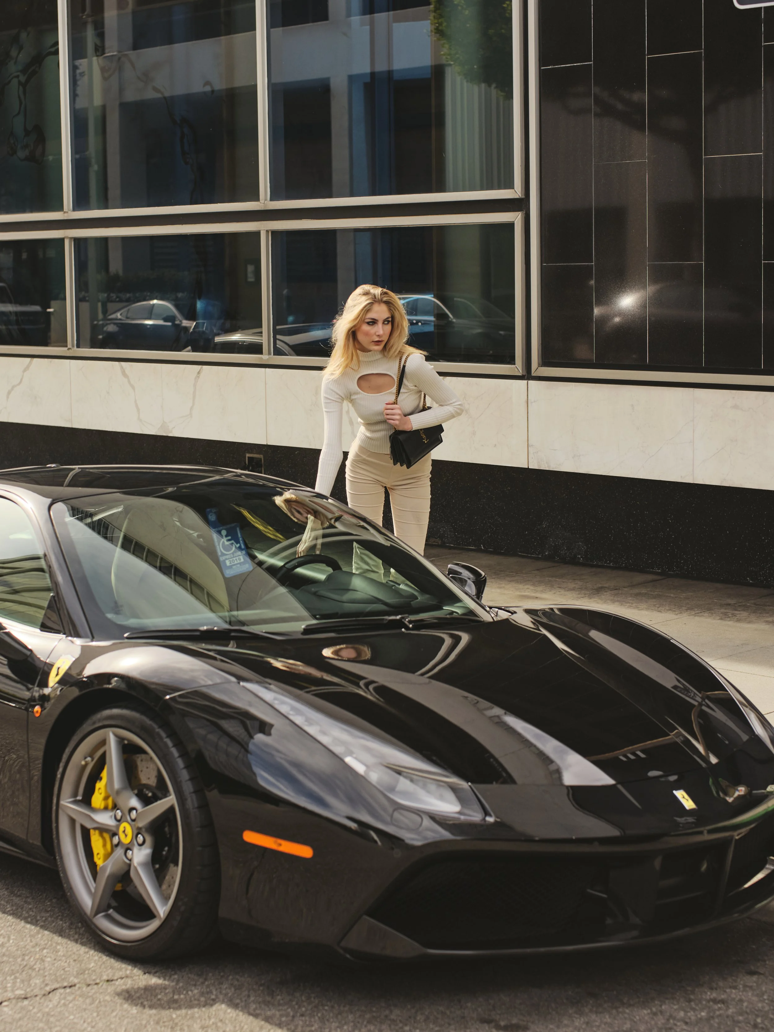 A woman with blonde hair, wearing a beige outfit with cut-out details, stands beside a black luxury sports car parked on the street in front of a modern glass building, holding a black handbag.