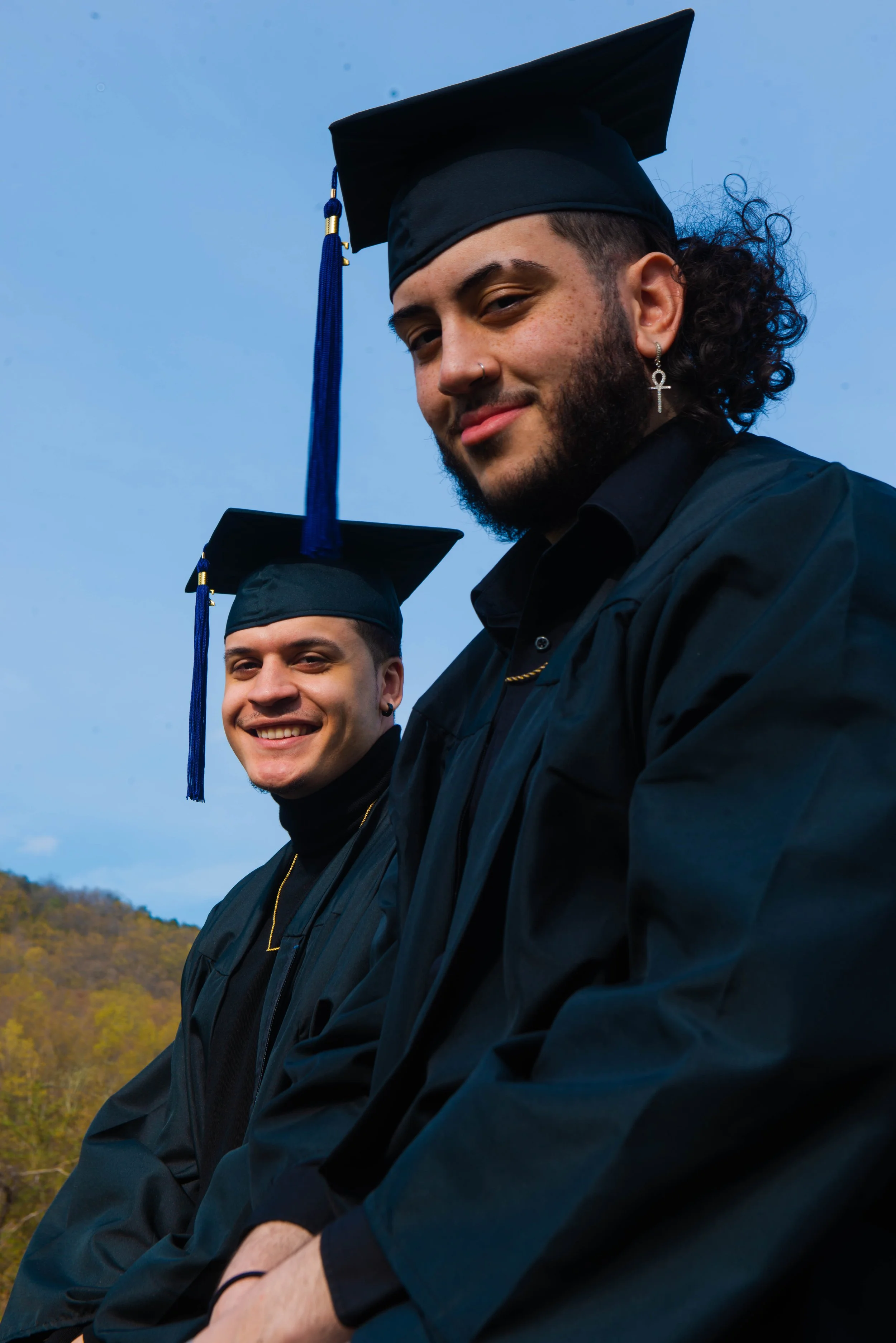 Two male graduates wearing black caps and gowns pose outdoors against a blue sky and distant trees.