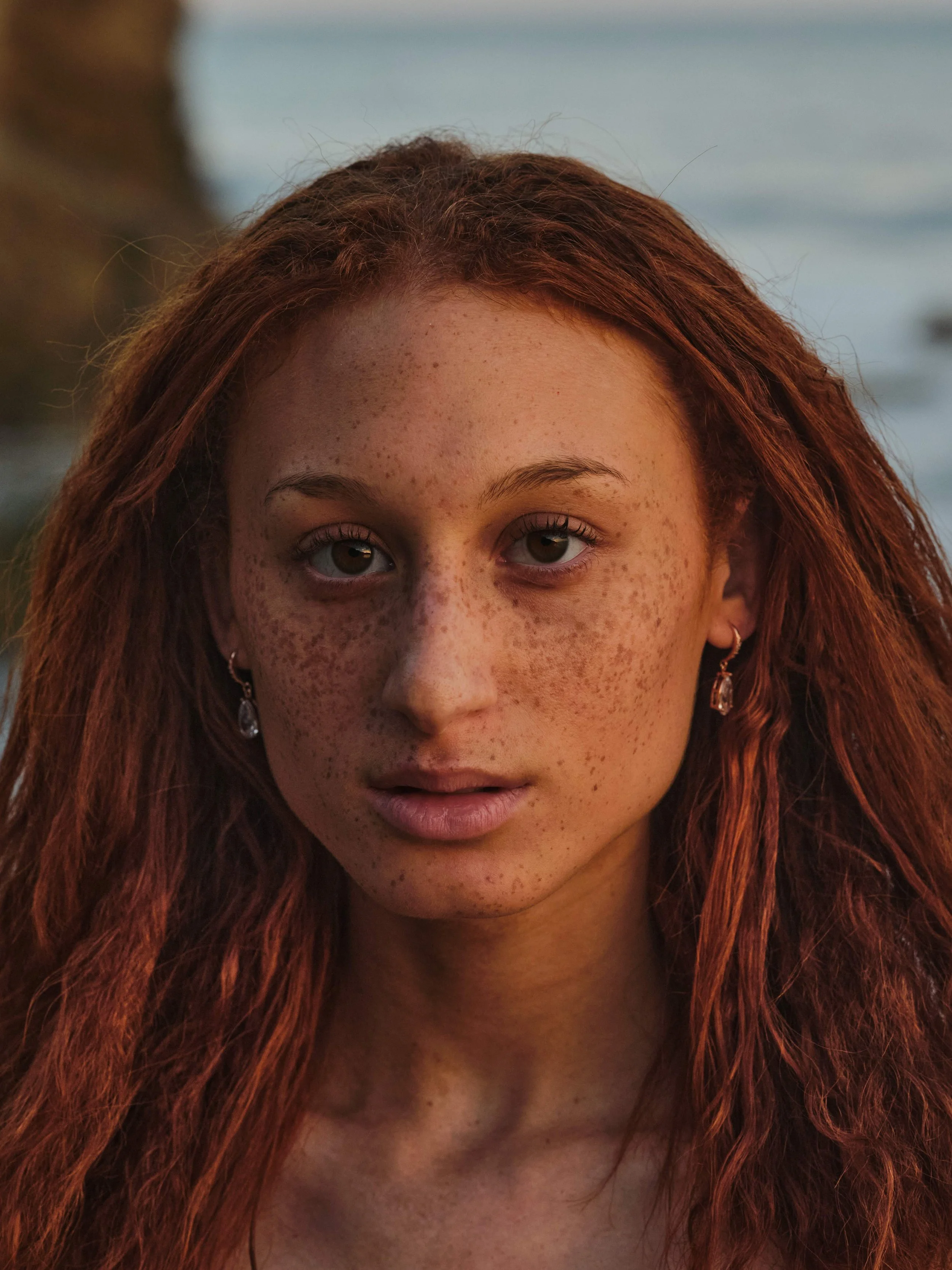 A young woman with red hair, freckles, and earrings looking directly at the camera outdoors near a body of water.
