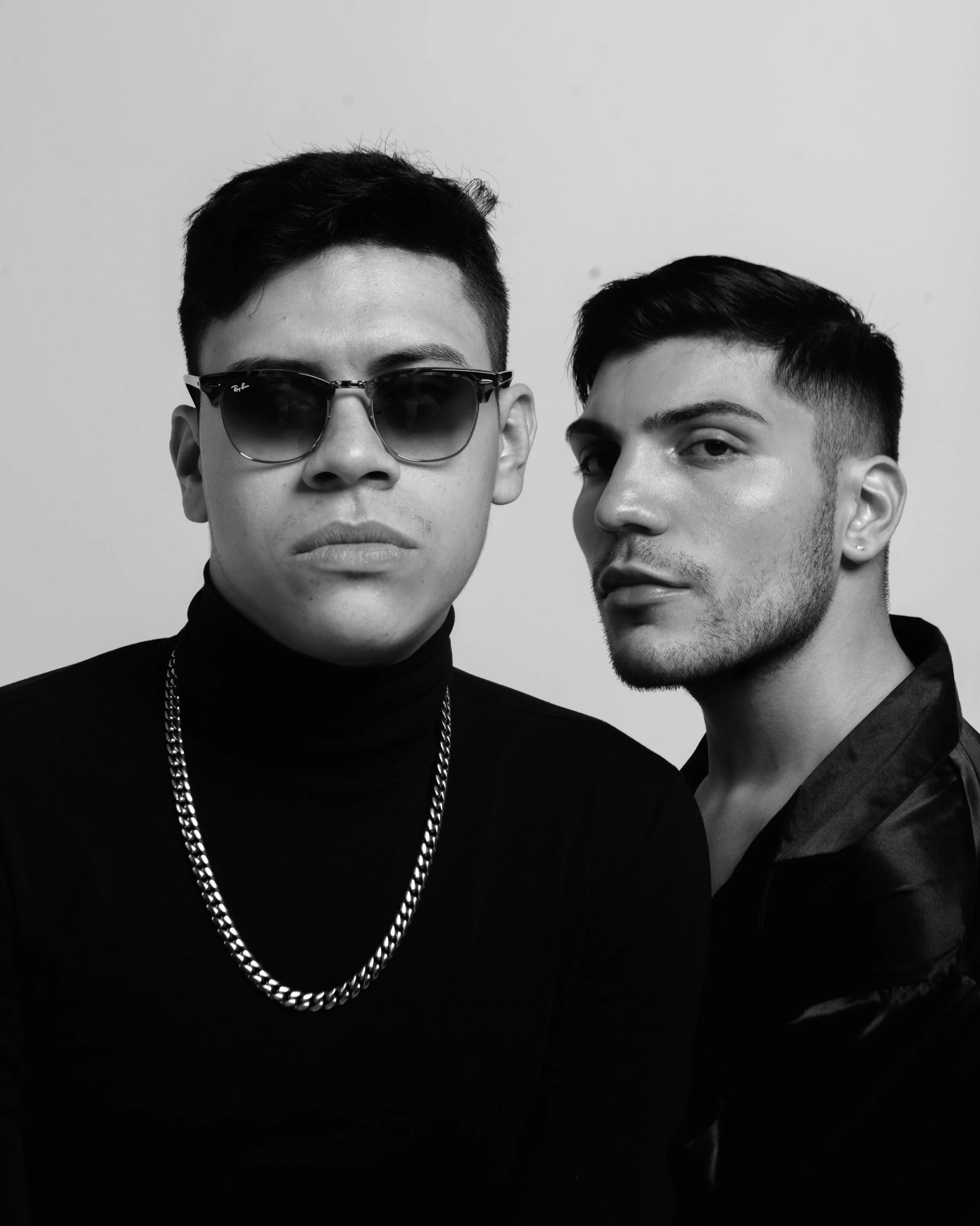 Black and white photo of two young men, one wearing sunglasses and a chain necklace, and the other without accessories, both with short hair and dressed in dark clothing, standing close together against a plain background.