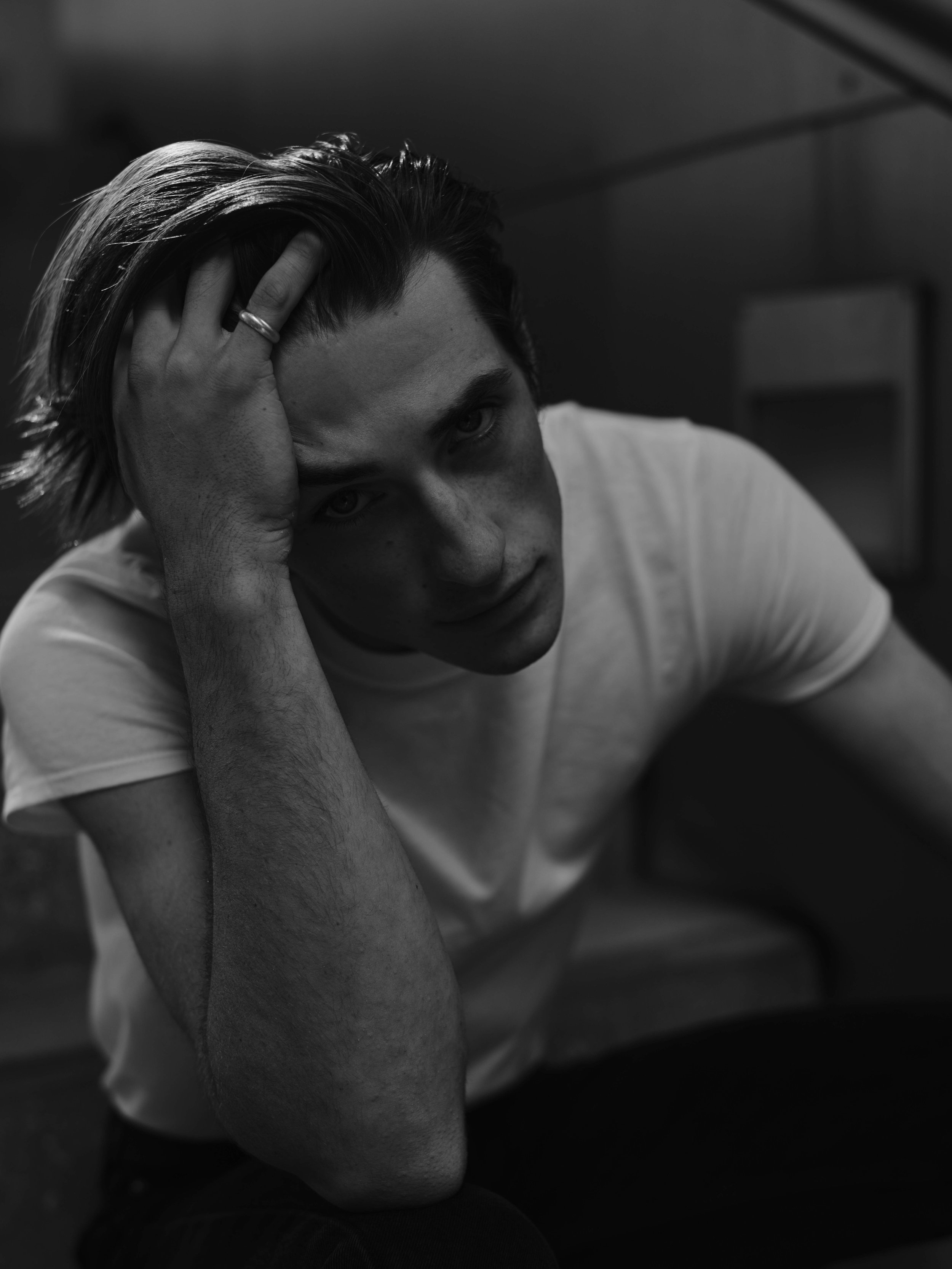 Black and white photo of a young man with dark hair, wearing a white t-shirt, sitting with his head resting on his hand, looking directly at the camera.