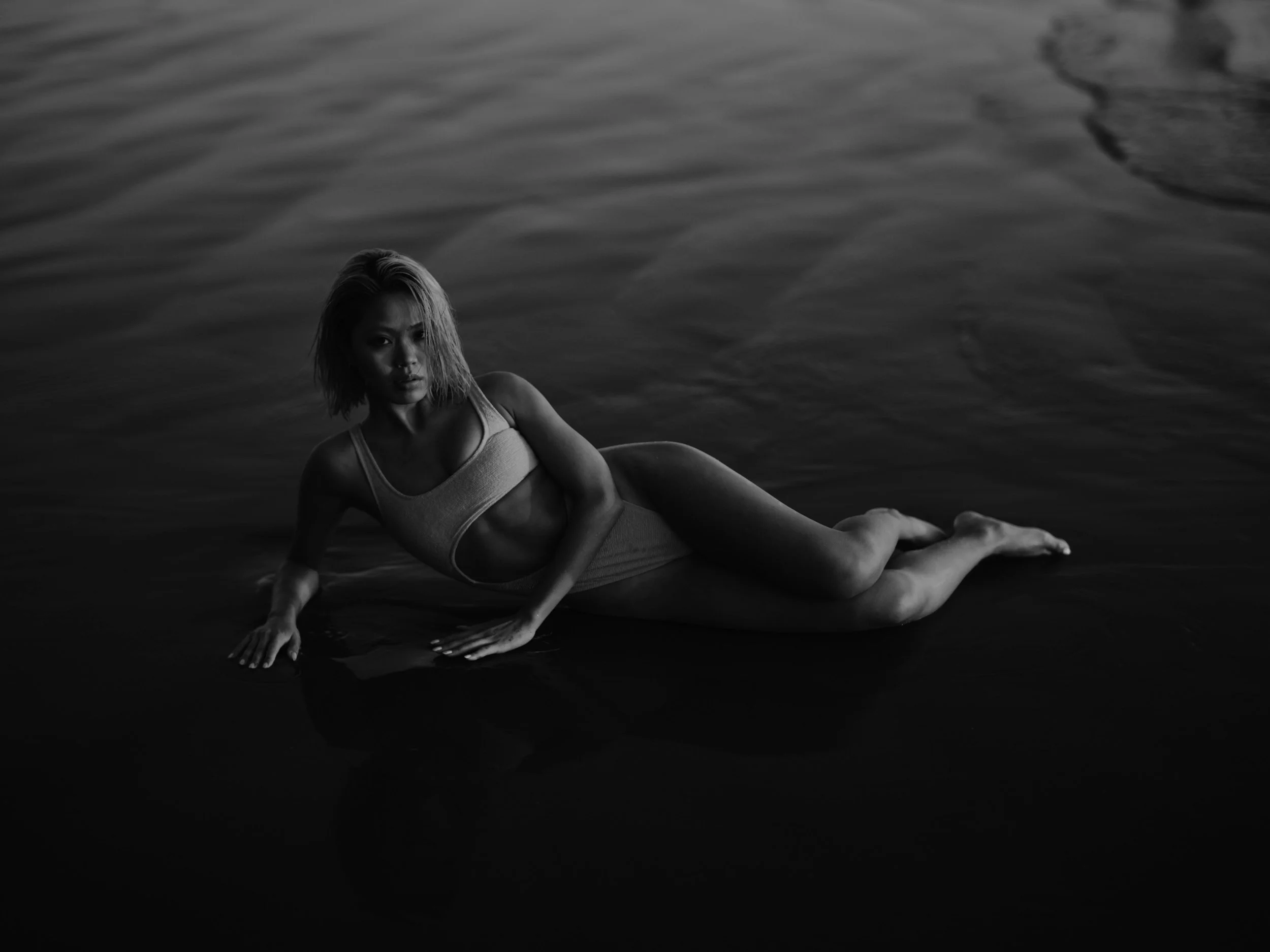 A woman in a swimsuit lying on her side in shallow water, with her hand touching the surface, in a black and white photo.