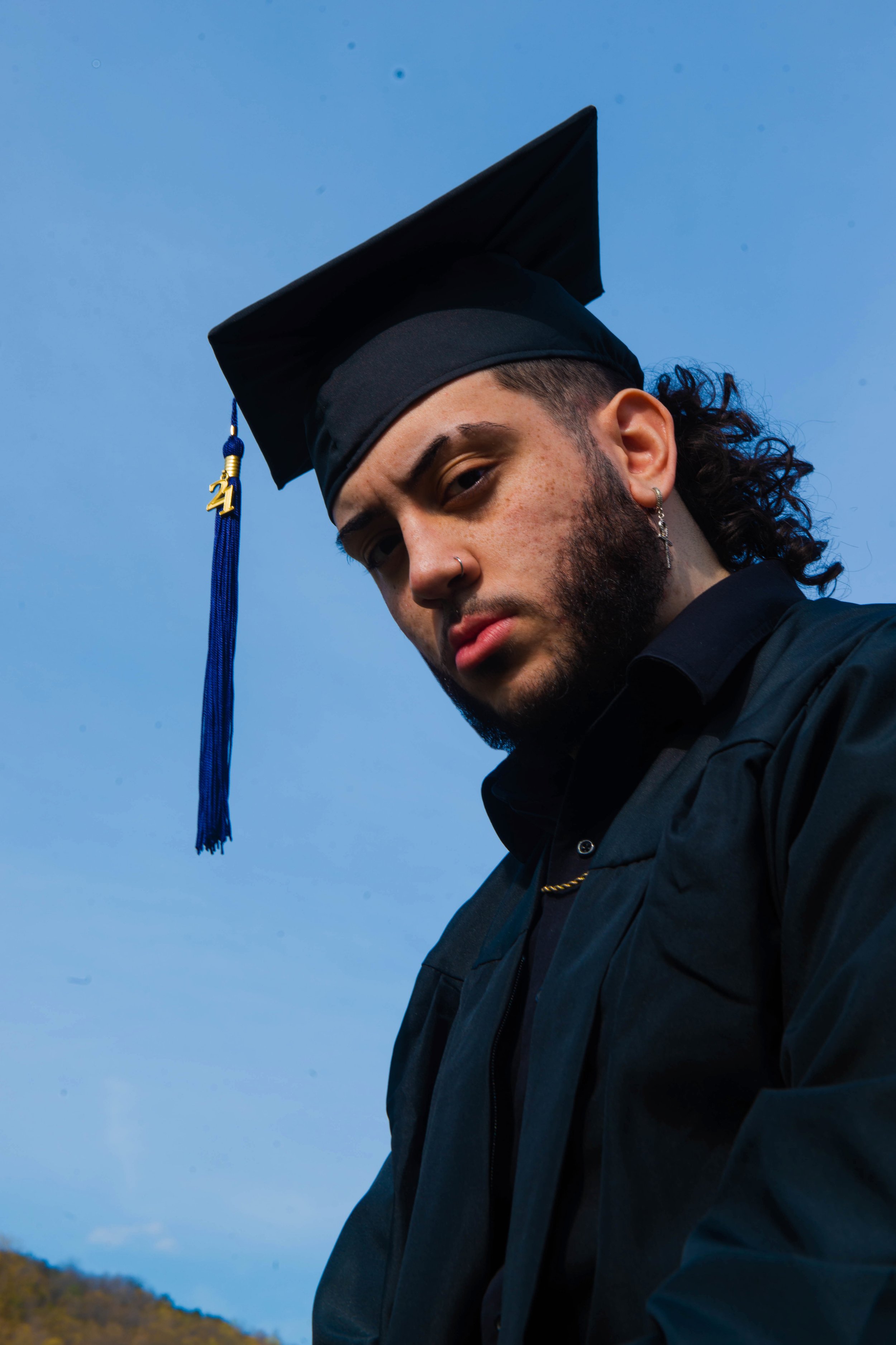 A young man wearing a graduation cap and gown stands outdoors with a blue sky in the background.