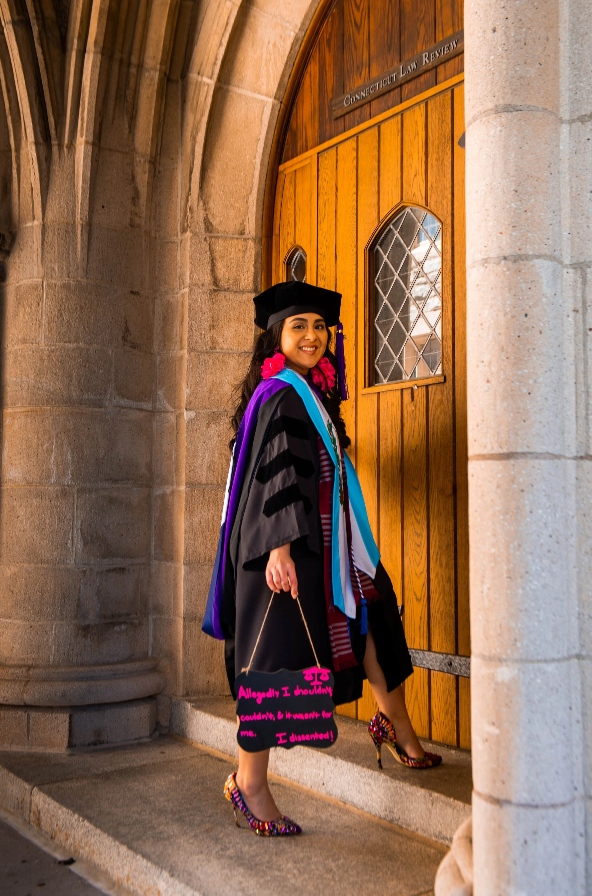 Woman in graduation attire standing outside a wooden door at a university