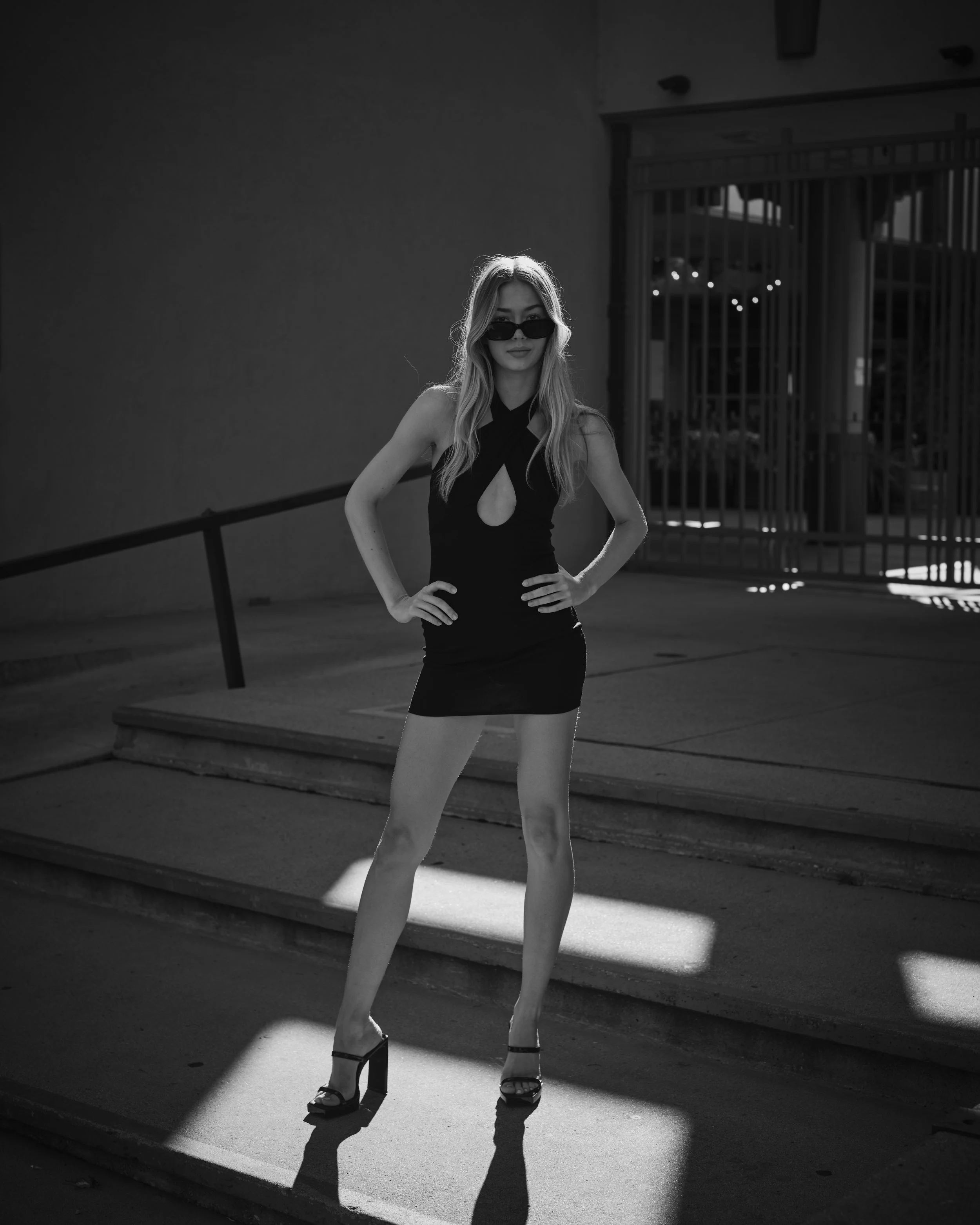 A woman wearing sunglasses and a short black dress with a cut-out design, standing on stairs with her hands on her hips in a shadowed outdoor area.