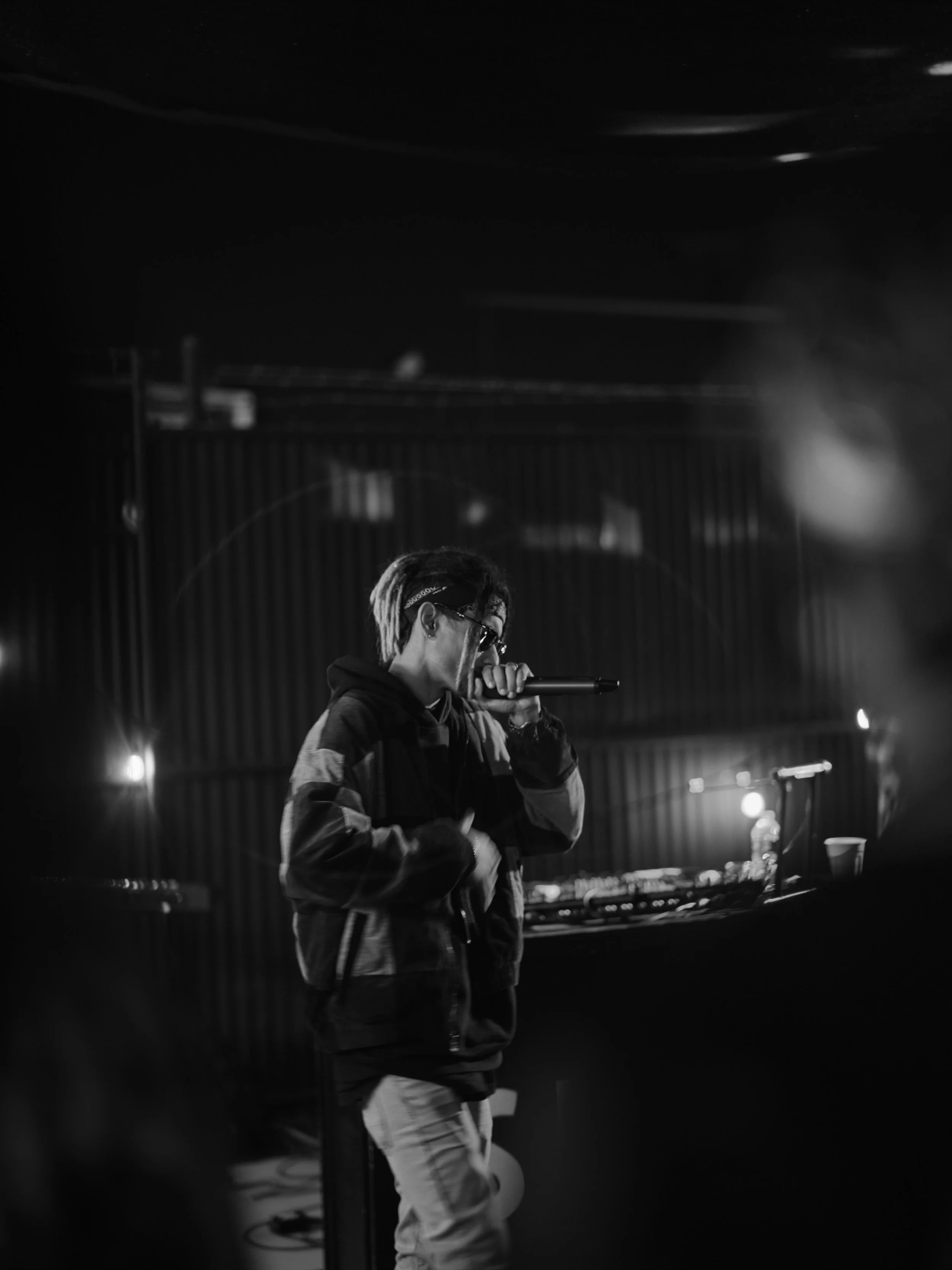 A person performing on stage with a microphone in black and white, wearing sunglasses and a hoodie, behind DJ equipment.
