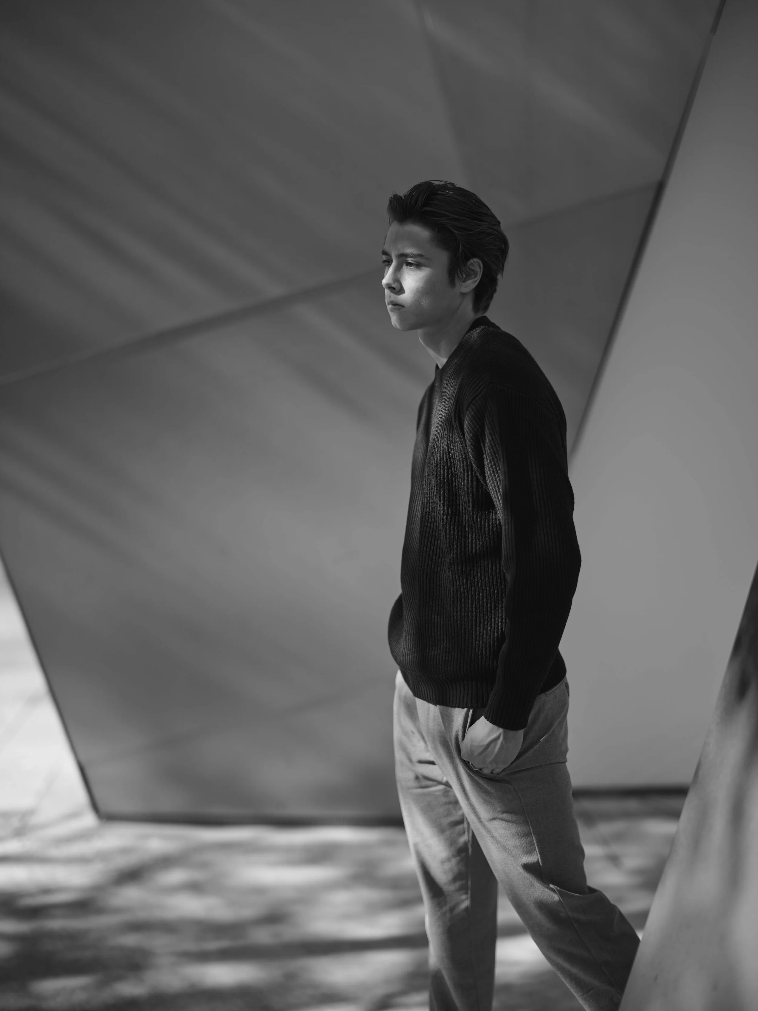 A black-and-white photo of a young man standing with his hands in his pockets, looking pensively to the left, against modern architectural background.