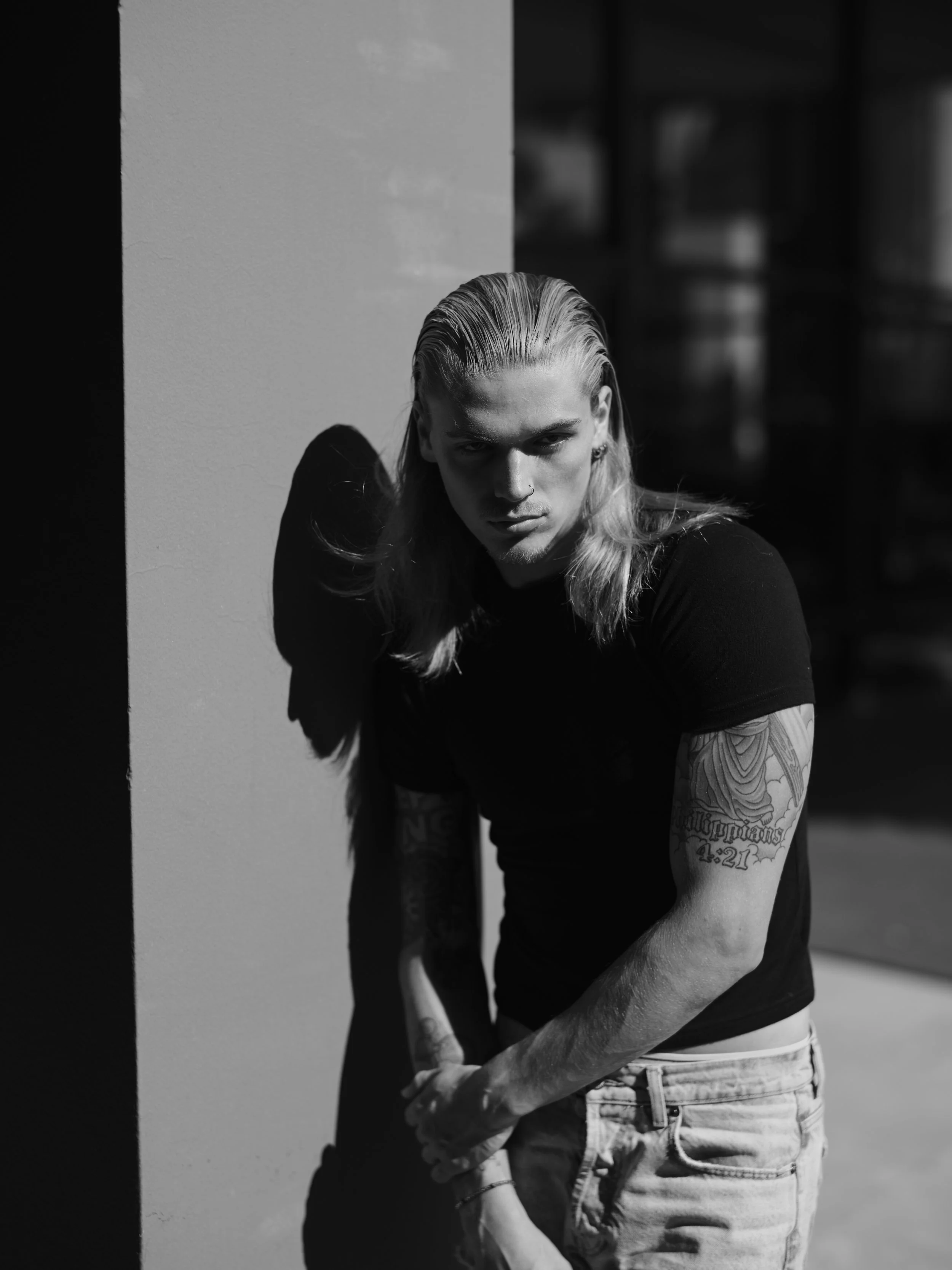 A black-and-white photo of a young man with long, slicked-back hair, standing against a wall with shadowed side, wearing a black t-shirt and light-colored jeans, with tattoos on his arms.