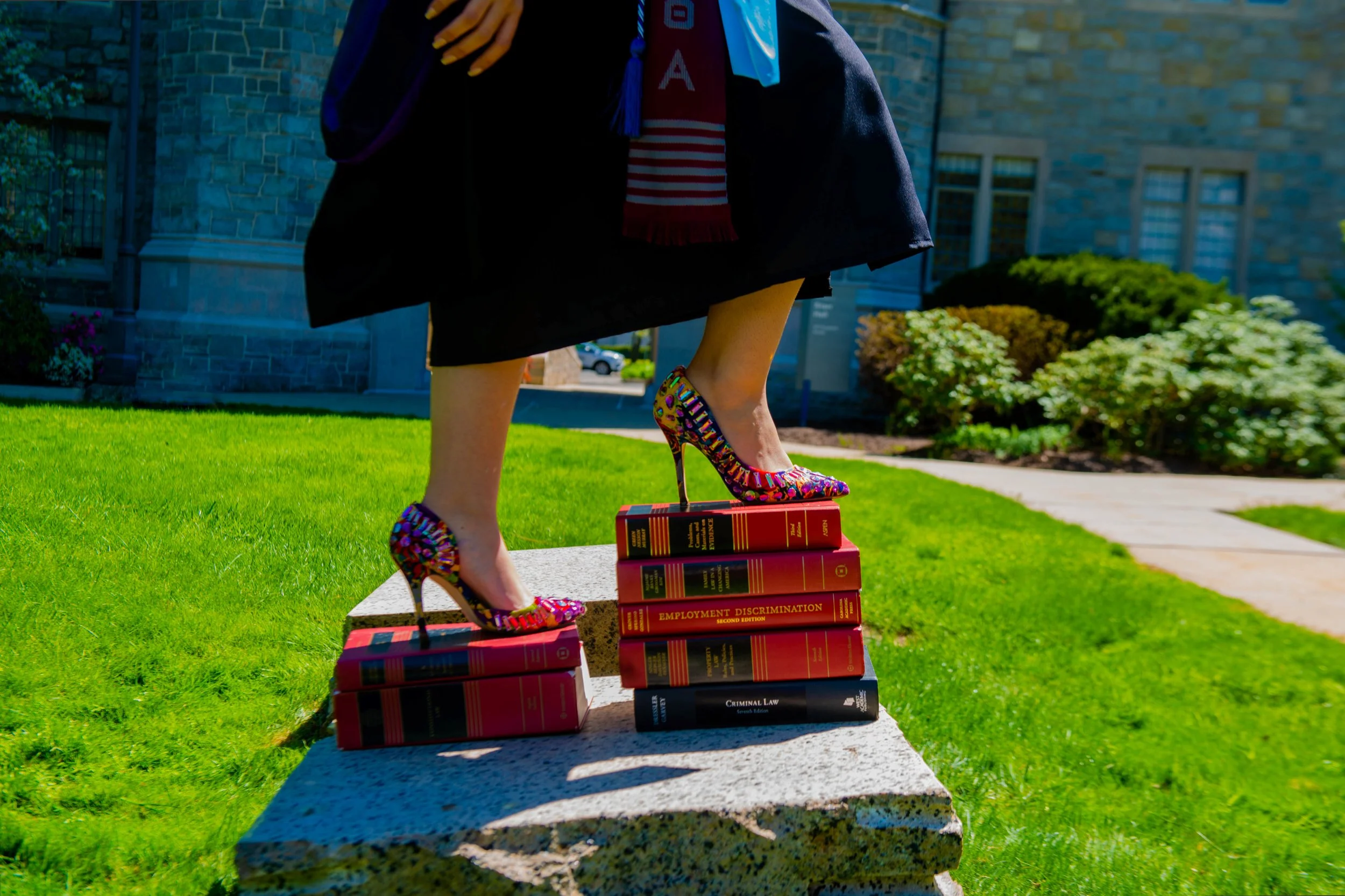 A person standing on a stone bench outdoors, wearing colorful high heels, a judge gown, and a graduation hood, with several law books stacked on the bench.