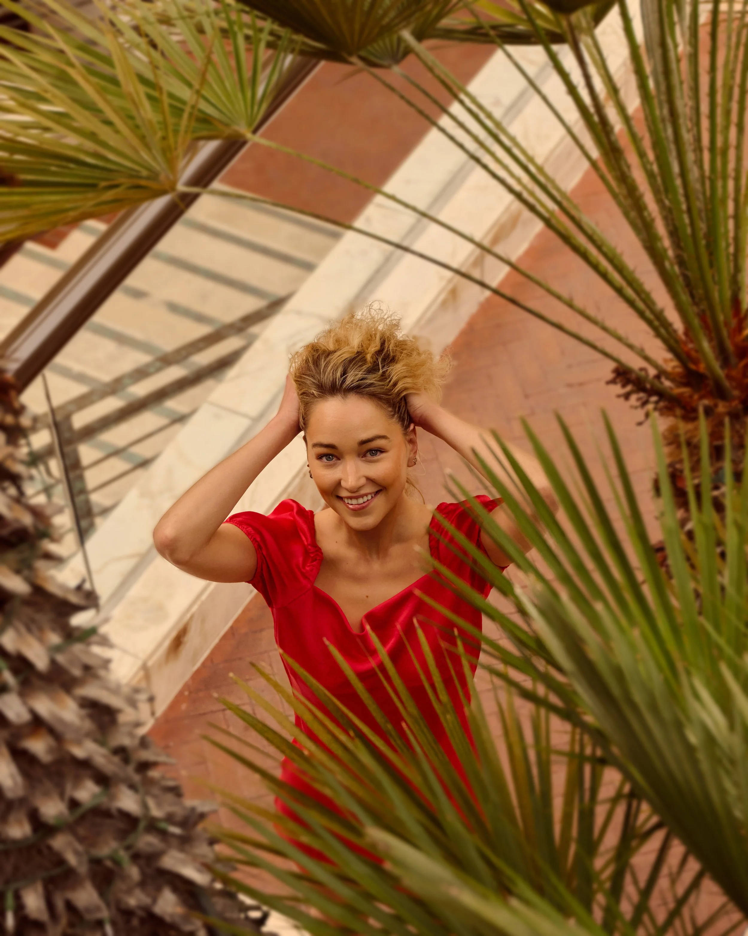 A smiling young woman in a red dress with tousled blonde hair, standing outdoors behind large palm leaves and looking up at the camera.
