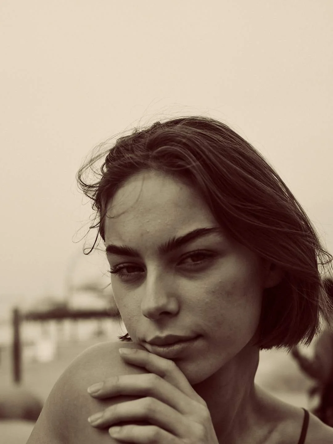Close-up black-and-white portrait of a young woman with short hair, gazing at the camera with a slight smile, resting her hand on her shoulder.