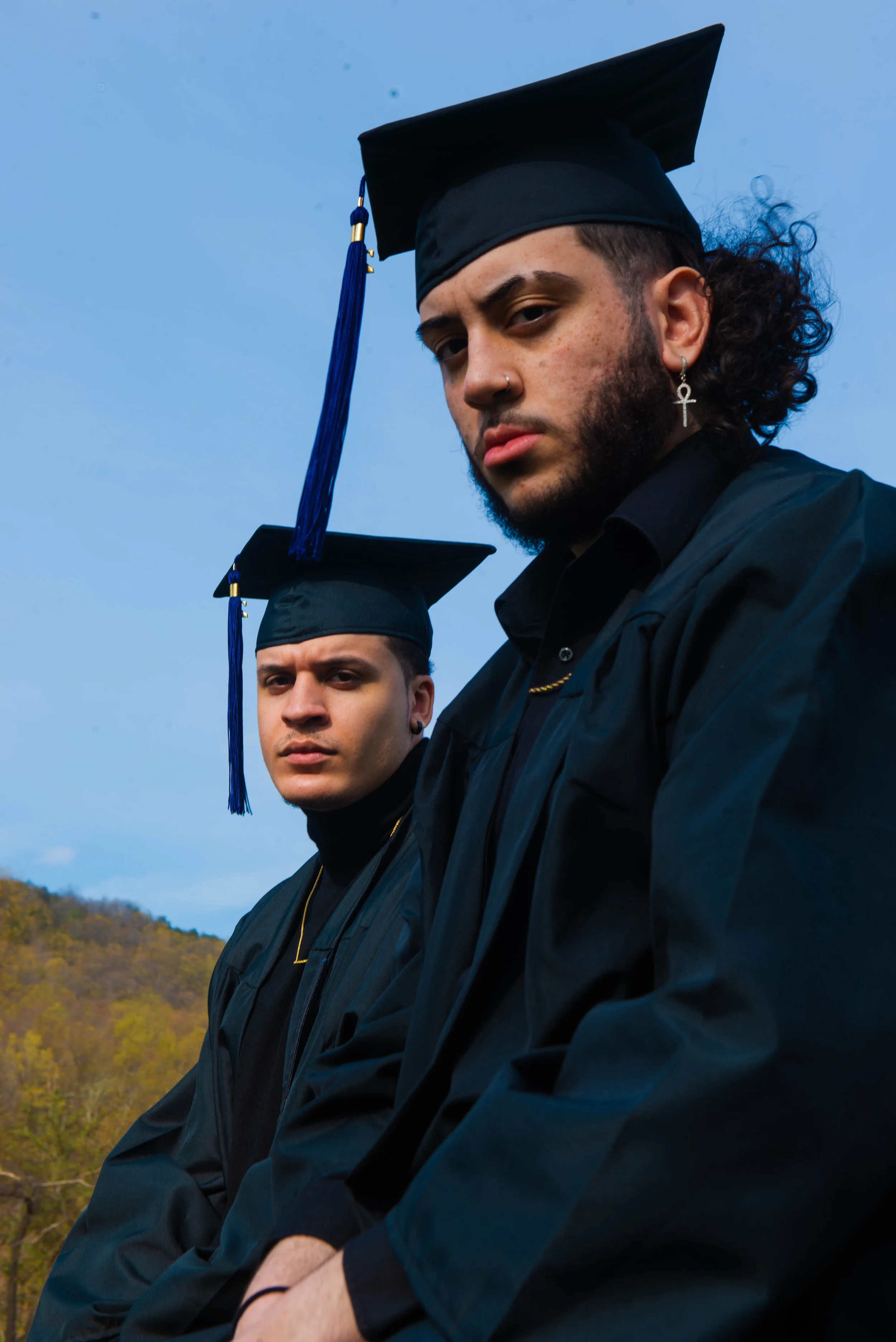 Two young men in graduation caps and gowns pose outdoors, with a blue sky and hillside in the background.