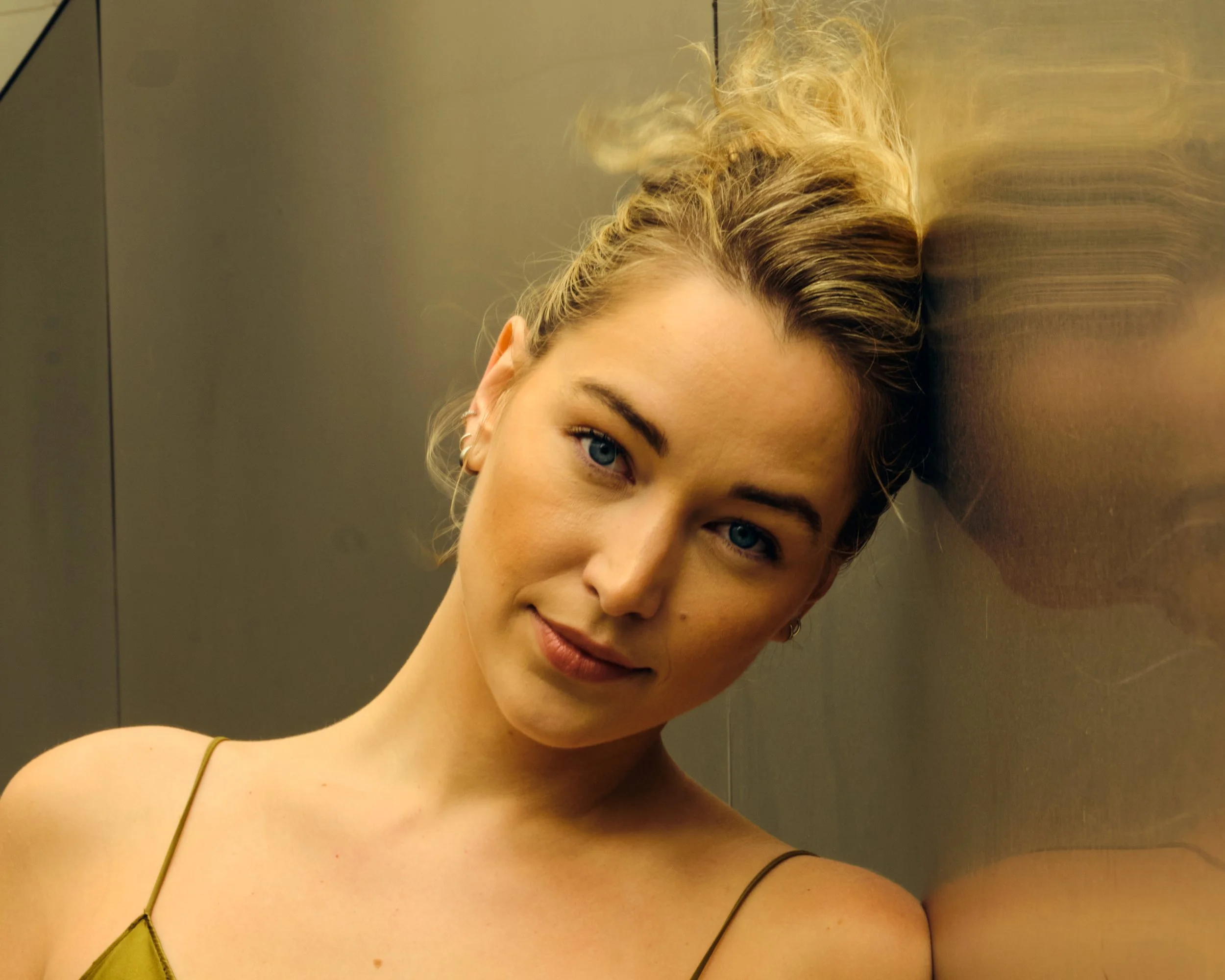 A woman with blonde hair and blue eyes leaning her head against a metallic wall, wearing a thin-strapped top.
